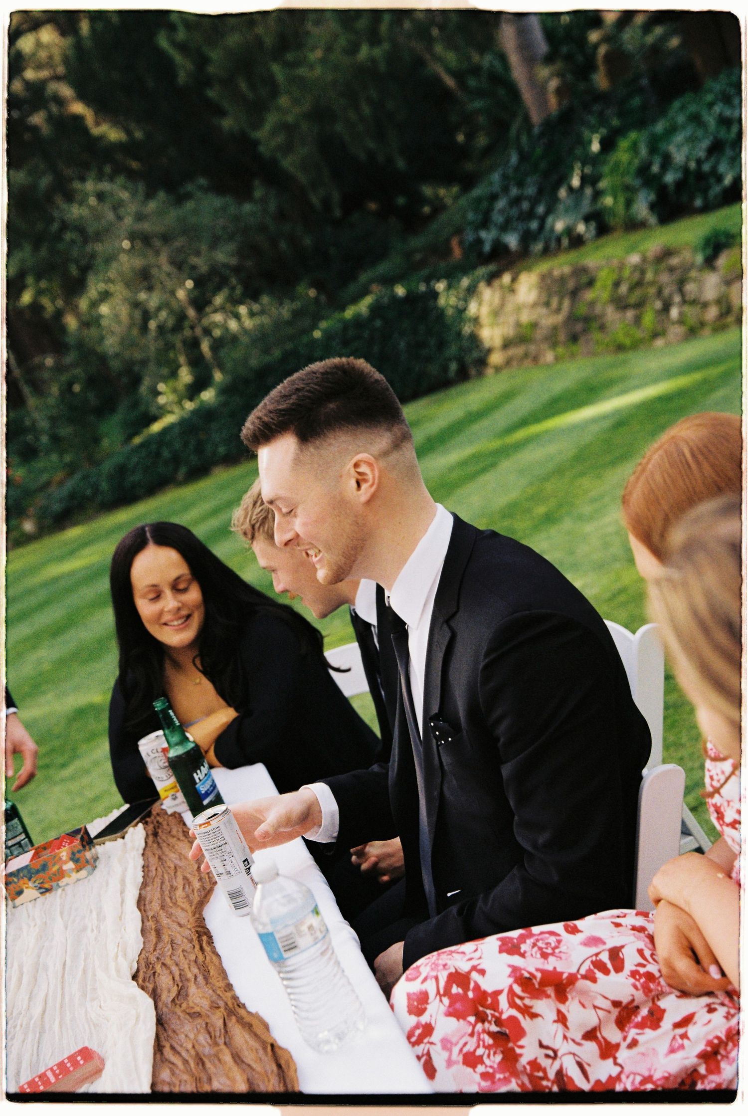 Groom in black tuxedo laughing with guests at outdoor wedding reception table on lush green lawn.