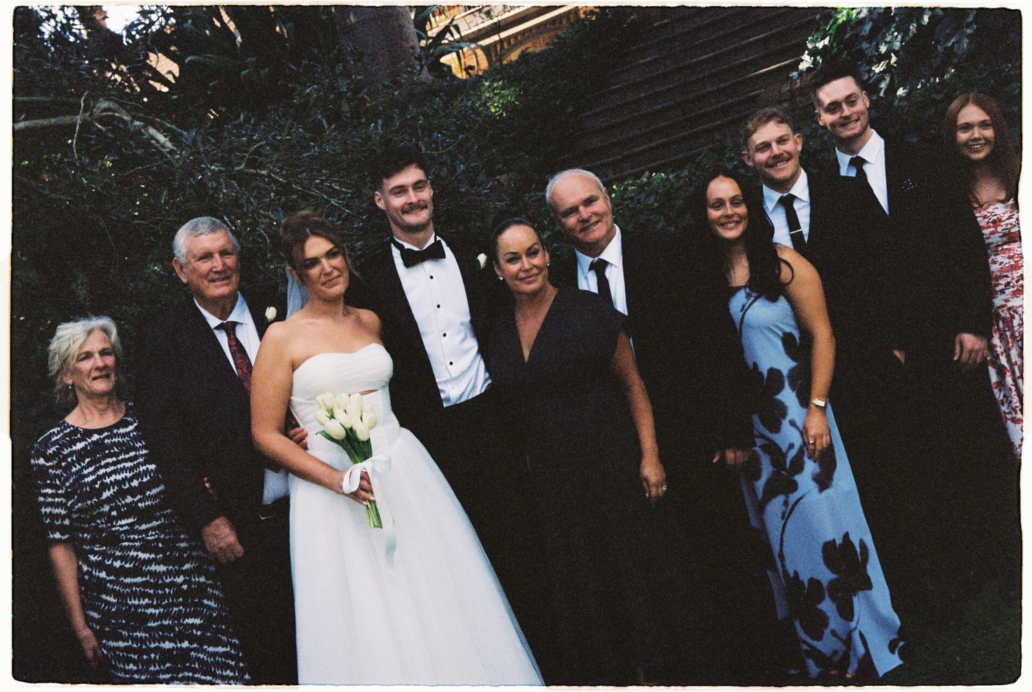 Wedding party group photo with bride in white gown and groom in tuxedo surrounded by family.