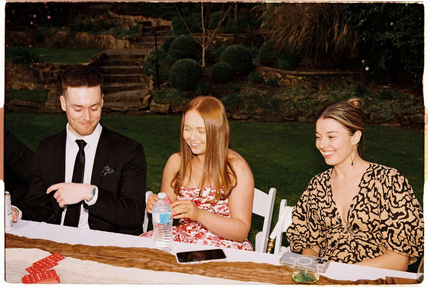 Three wedding guests seated at outdoor reception table, including a woman in floral dress.
