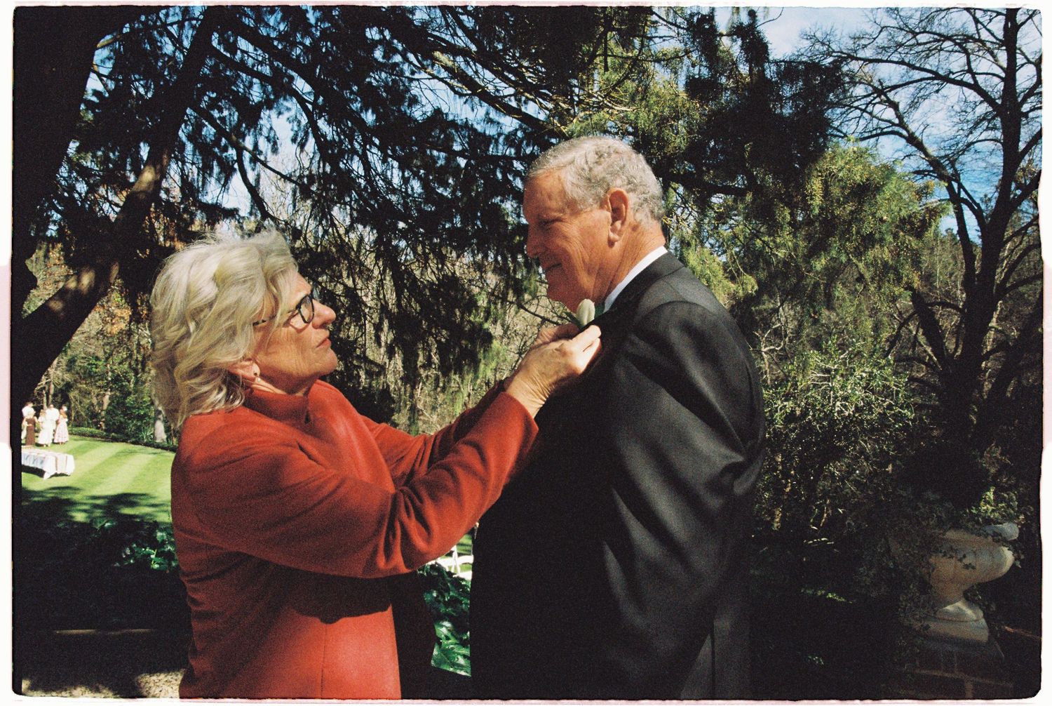 Woman in red jacket adjusting boutonniere on mans suit jacket outdoors under trees.