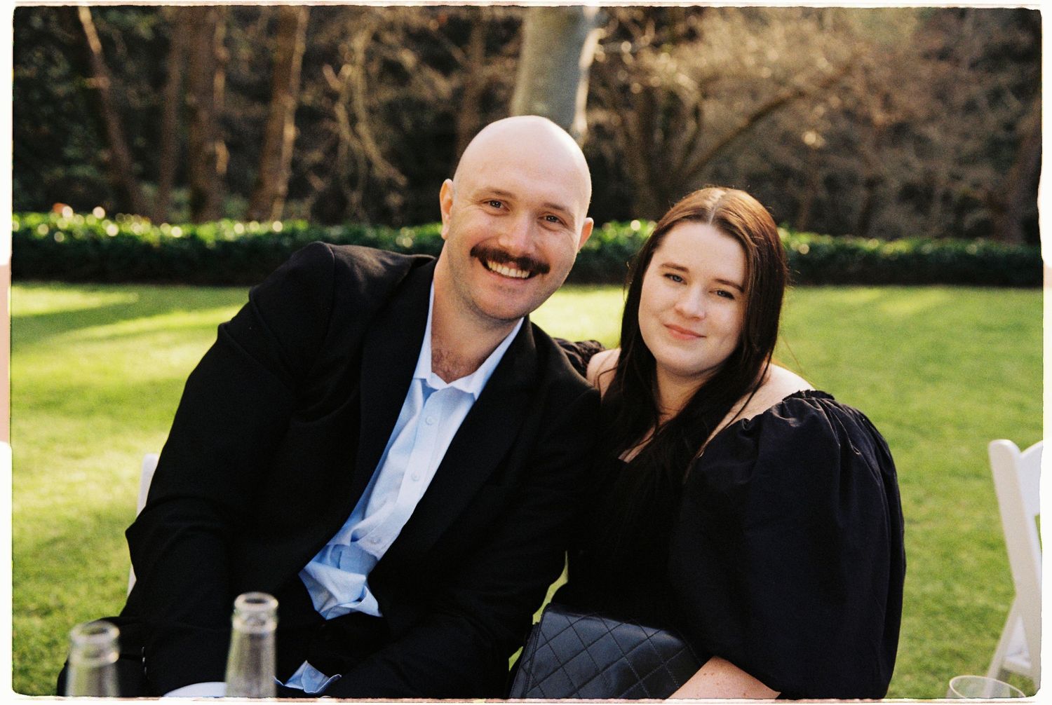 A bald man with a mustache and a young woman smile together at an outdoor garden event.