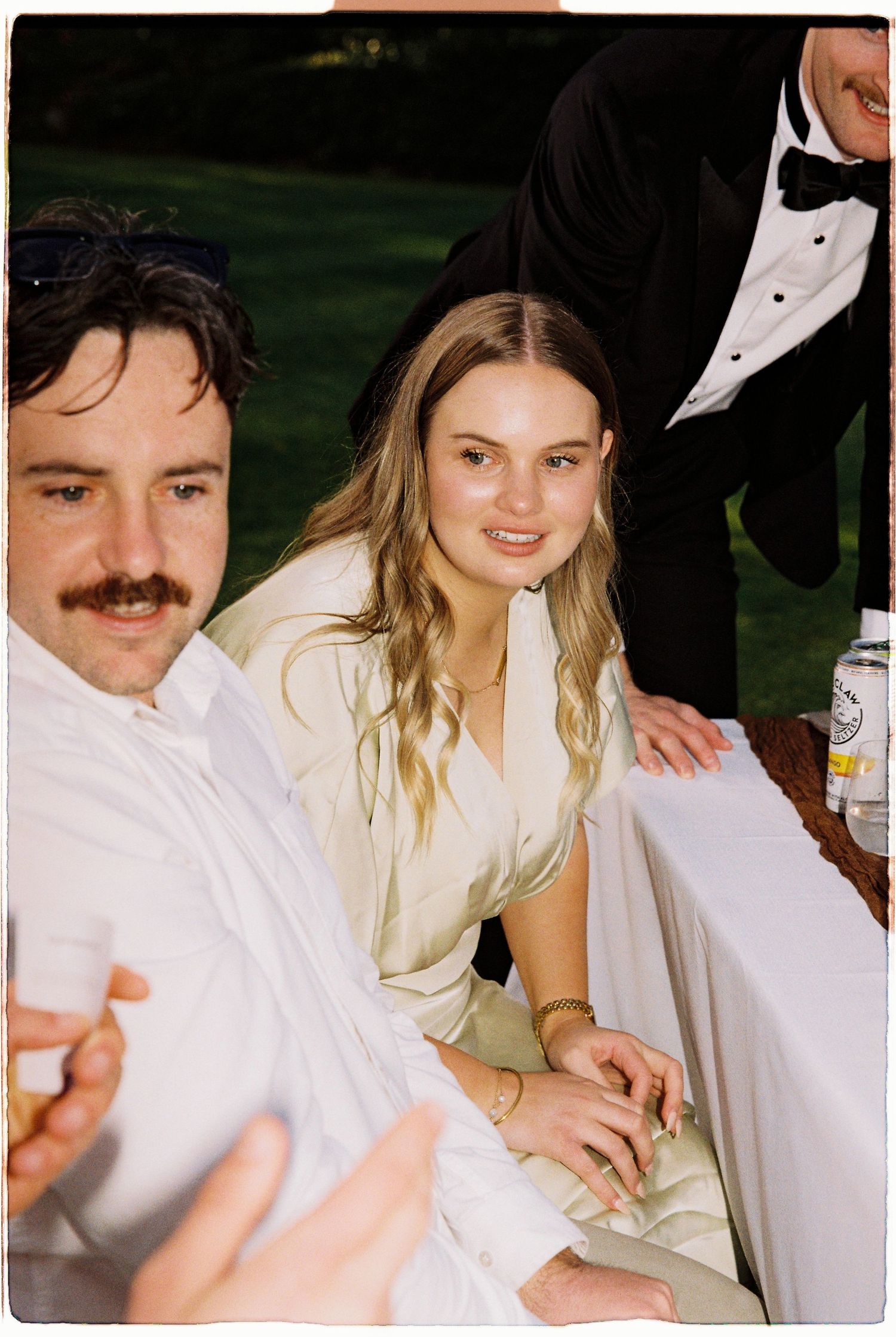 Man with mustache and blonde woman in sage dress seated together at an outdoor wedding reception.