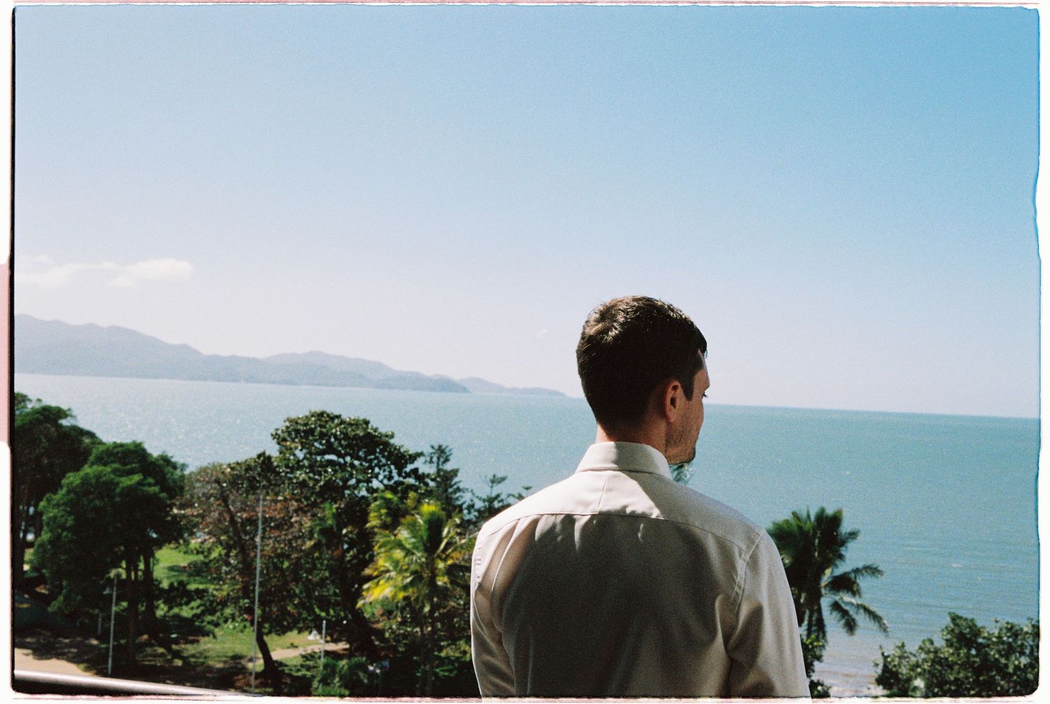 A man in a white shirt gazes at a scenic ocean view with lush tropical trees and mountains in the background.