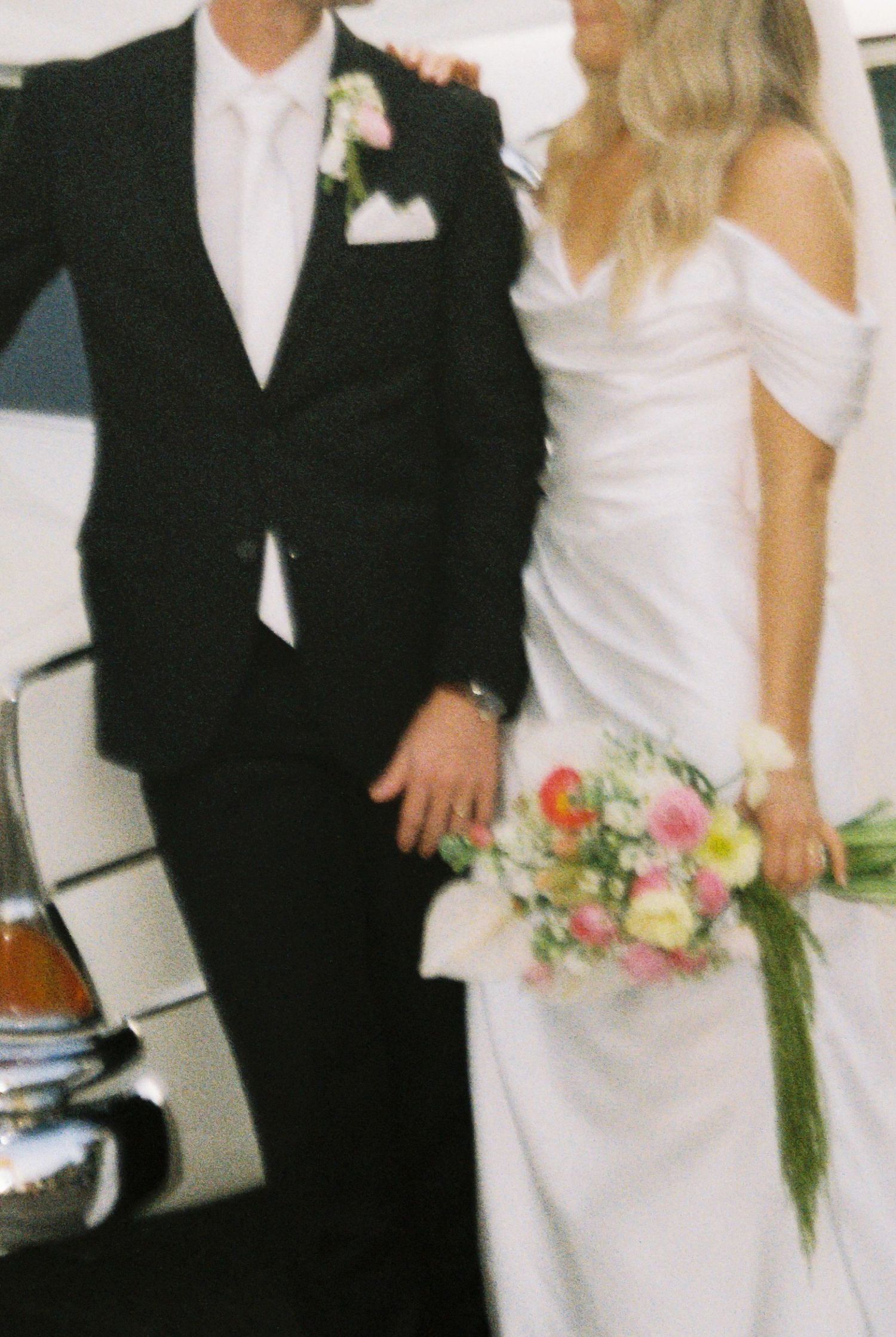Close-up of bride holding colorful spring bouquet in off-shoulder gown beside groom in black tuxedo.