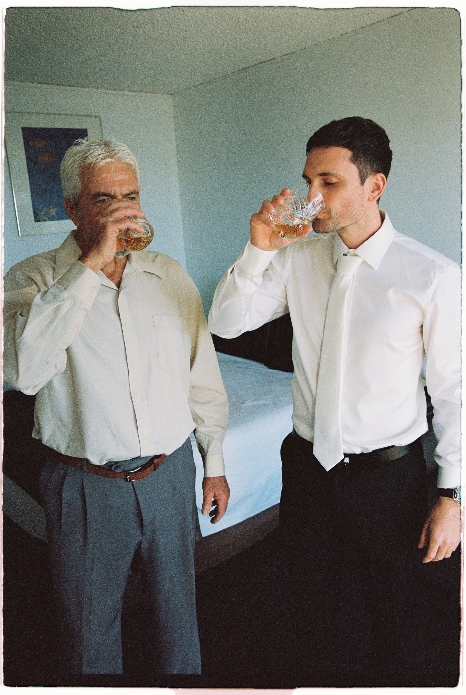 Two men in white dress shirts drinking whiskey from crystal glasses in a hotel room.