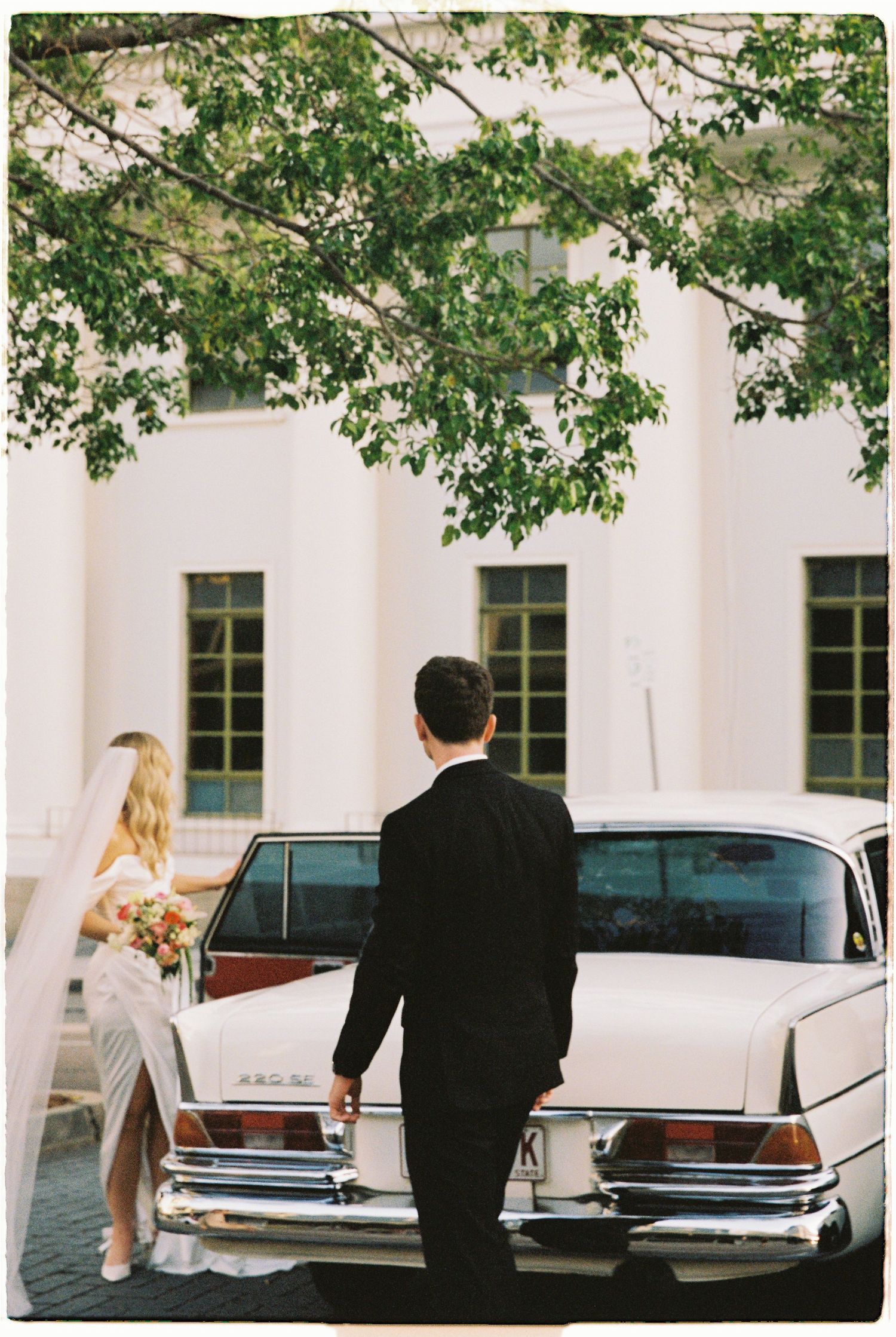 Bride and groom stand by a classic vintage white car outside a grand white building beneath a lush green tree canopy.
