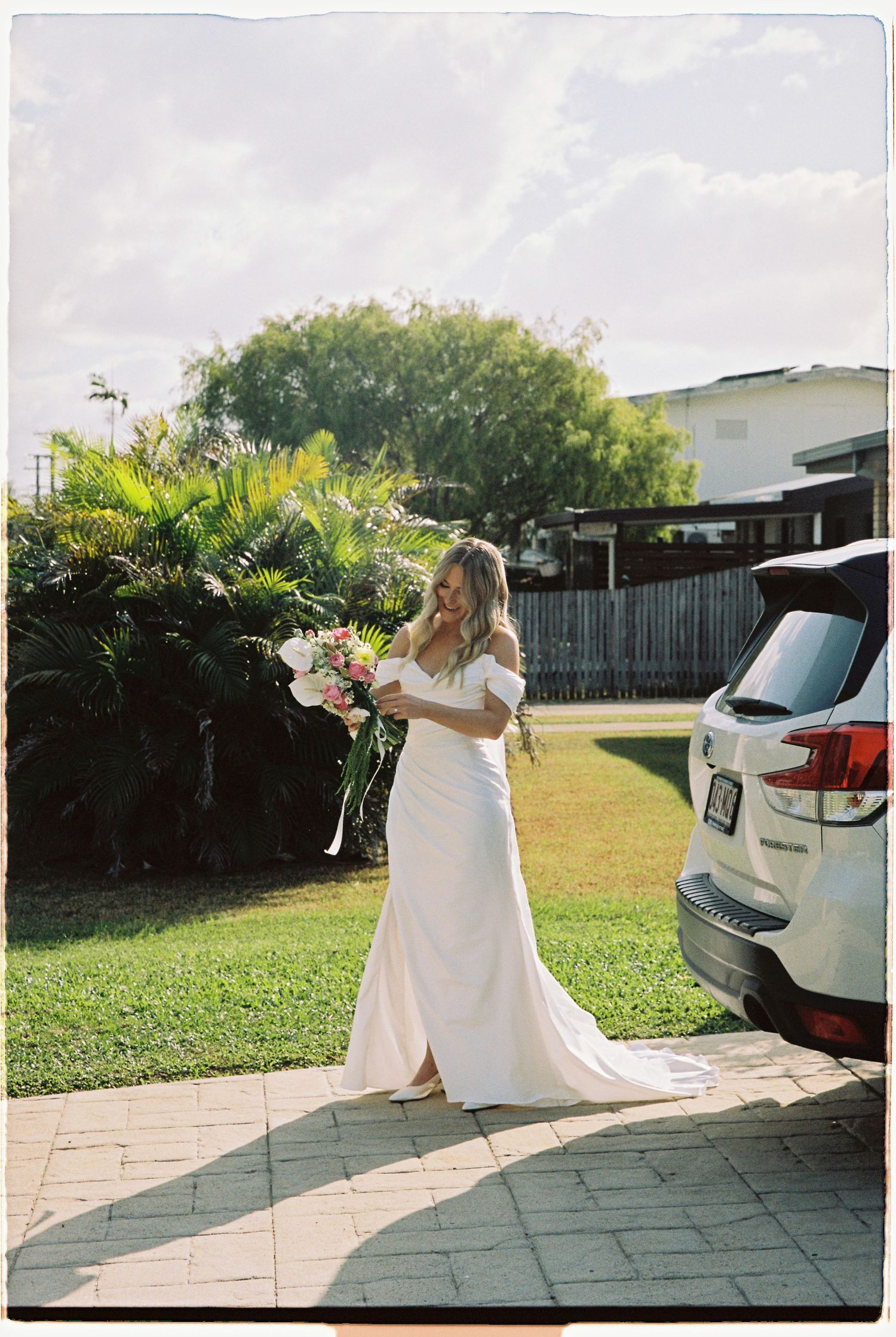 Bride in white off-shoulder gown holding pink bouquet beside car in sunny garden.