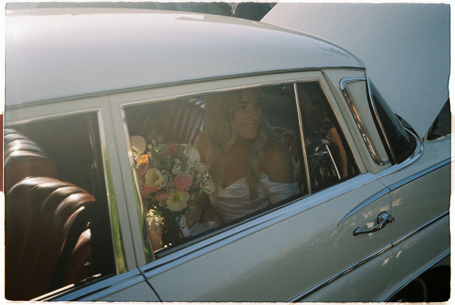 Bride with floral bouquet seated inside vintage silver wedding car in warm sunlight.