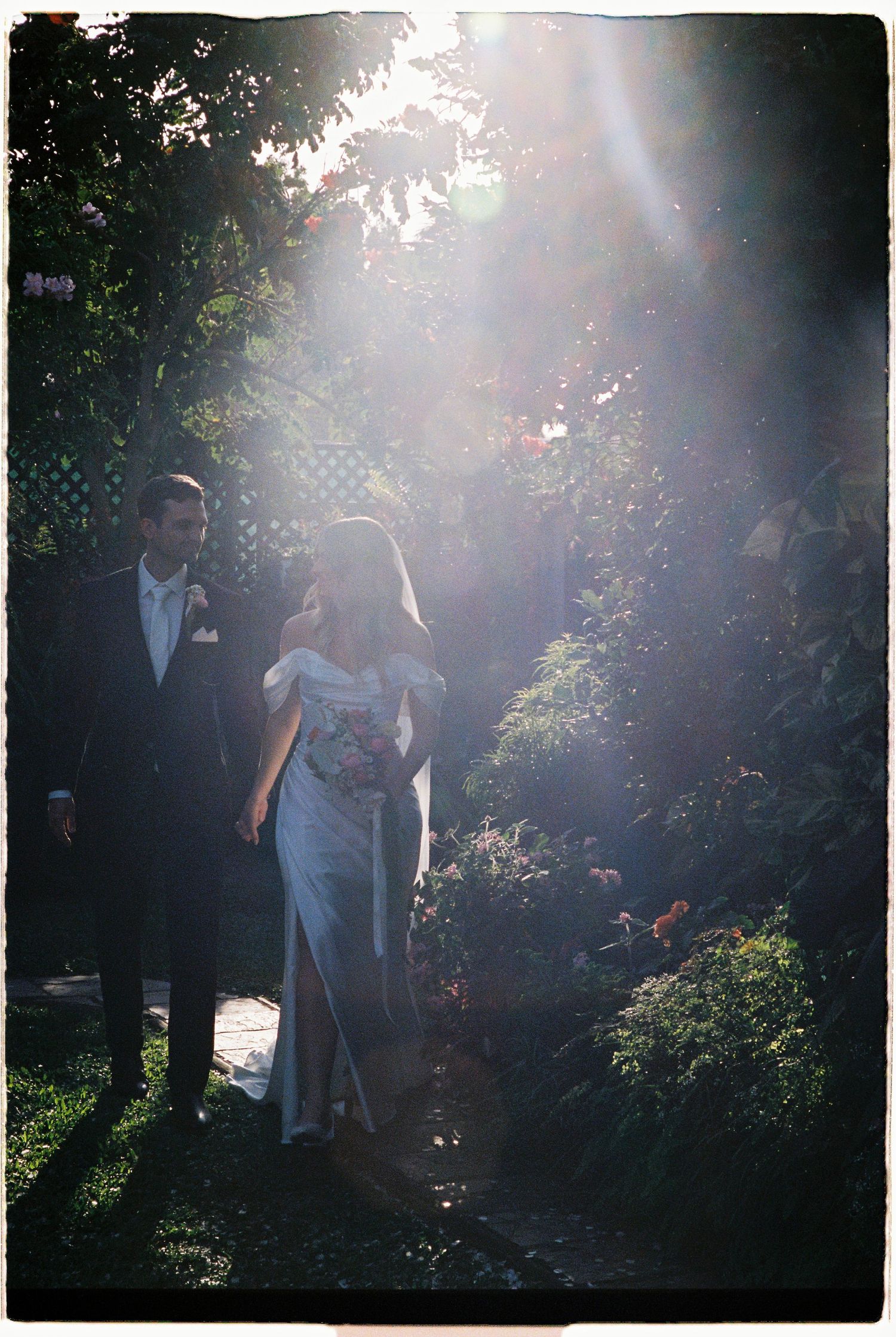 Bride and groom walking through sunlit garden with dreamy lens flare effect.