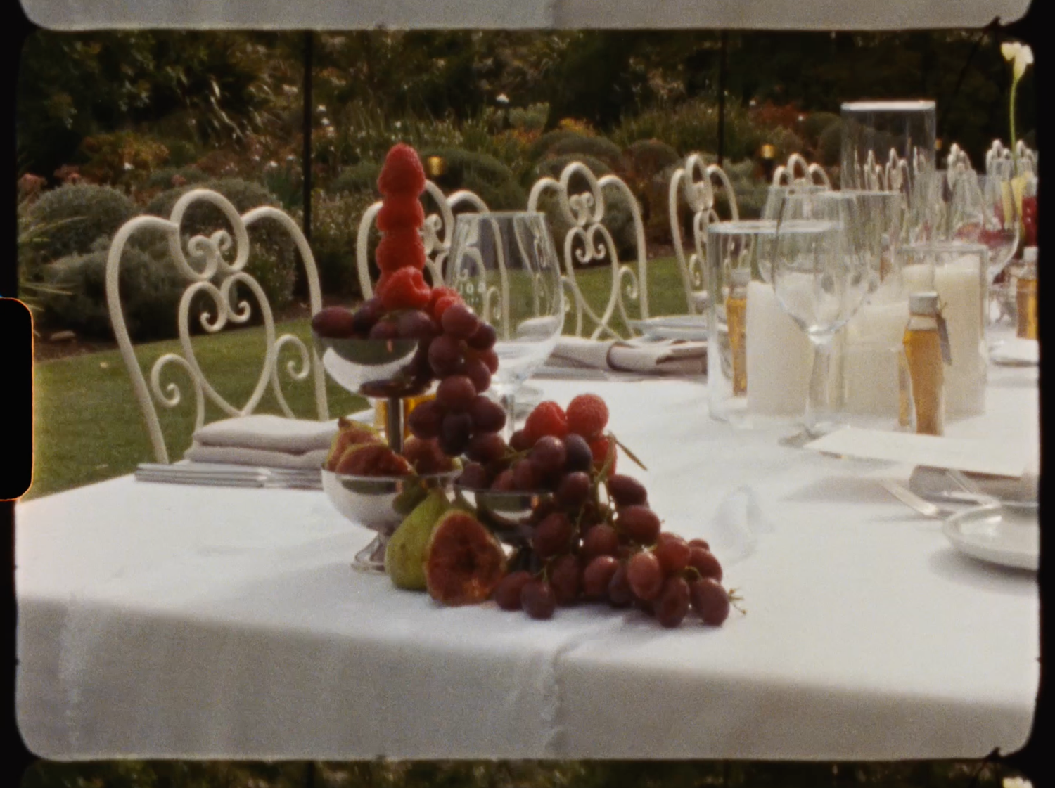 Elegant outdoor dining table with white tablecloth, heart-shaped chairs, fresh fruit display, and champagne glasses set for a garden party.