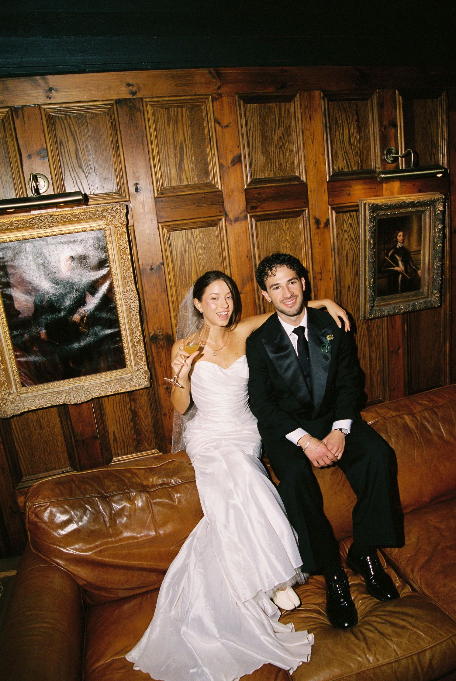 Bride and groom sitting together on a brown leather sofa in a wood-paneled room with framed paintings.