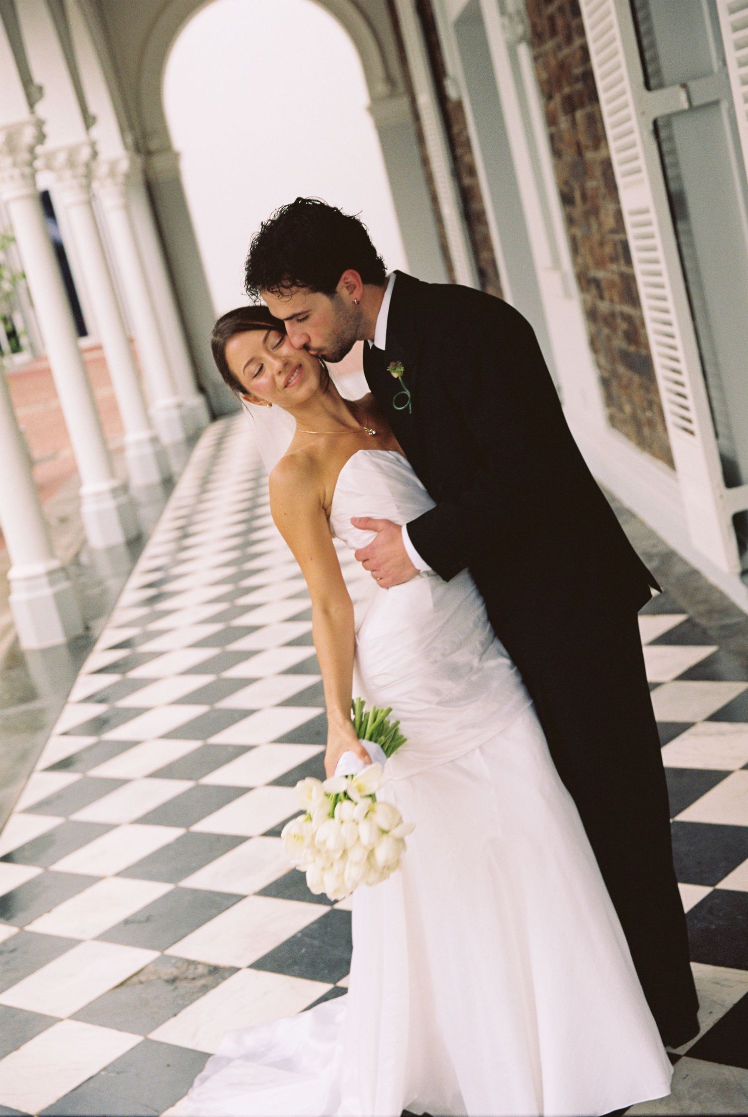 Bride in white gown holding bouquet embraced by groom in black tuxedo on checkered floor of elegant venue.