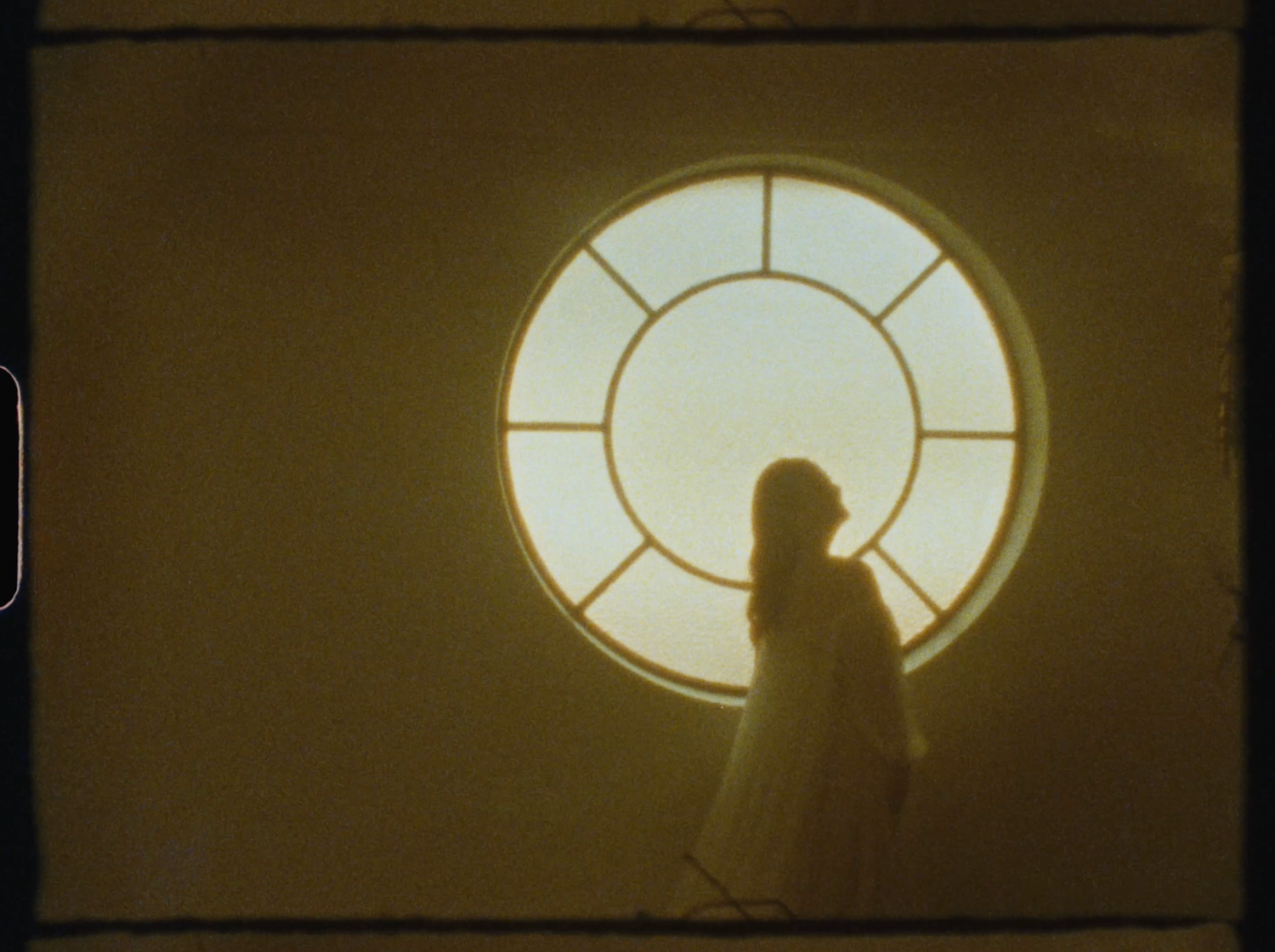 Silhouette of a woman standing before a large circular window with warm golden backlight in a dreamy, cinematic setting.