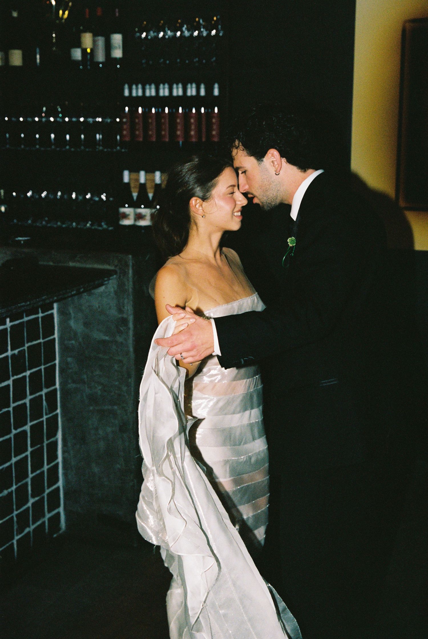 Bride in strapless mermaid gown and groom embrace tenderly near wine display at wedding venue.