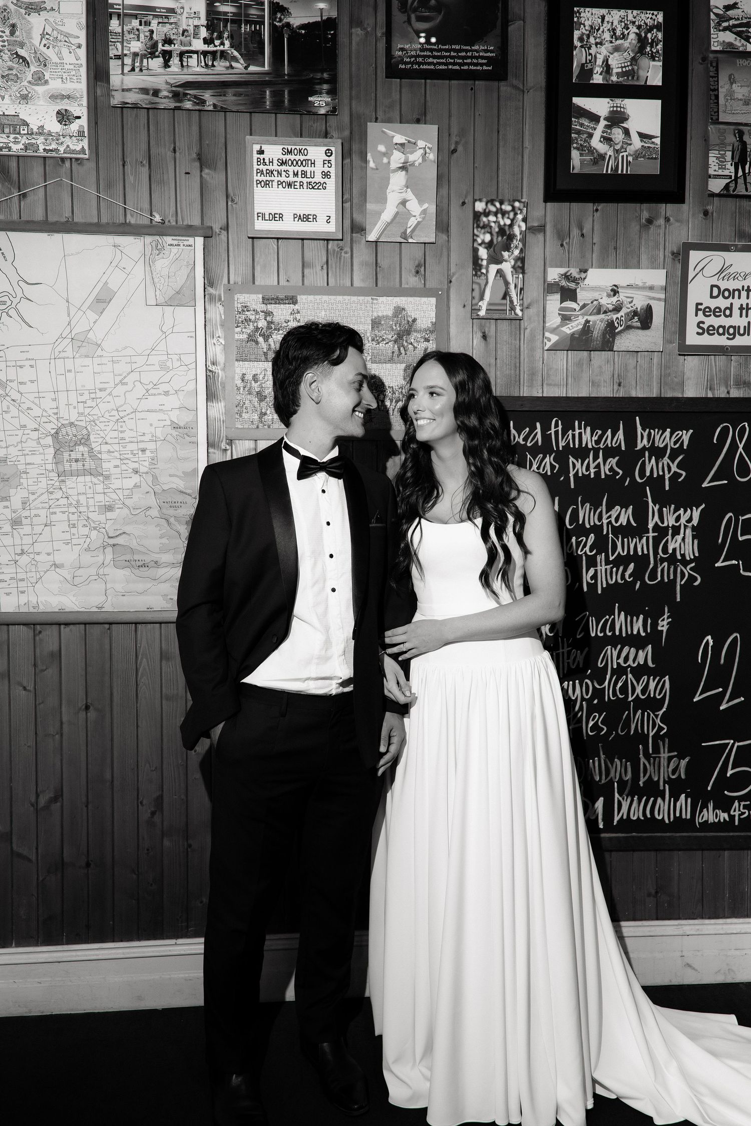 Bride and groom pose together in black and white inside a rustic restaurant with a chalkboard menu backdrop.