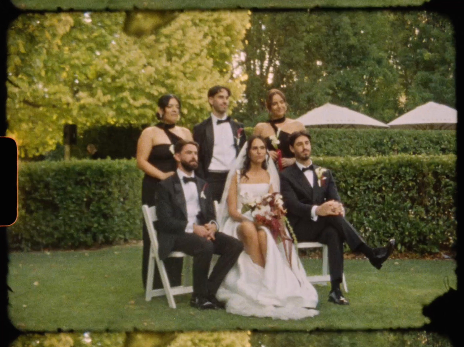 Wedding party group photo on green lawn with bride, groom, bridesmaids and groomsmen in formal attire.