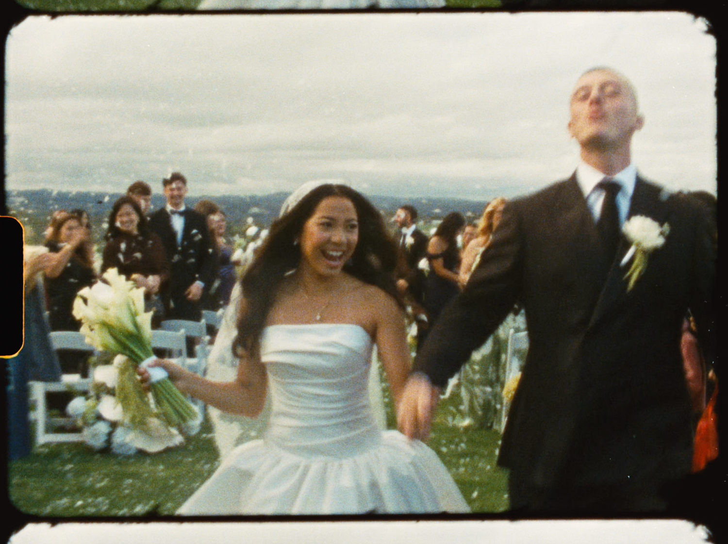 Radiant bride laughing with bouquet as groom walks beside her after outdoor wedding ceremony.