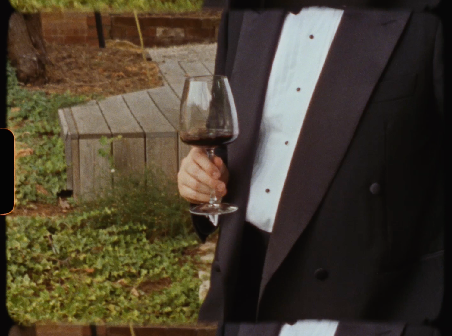 A person in a tuxedo holding a glass of red wine outdoors at a wedding reception.