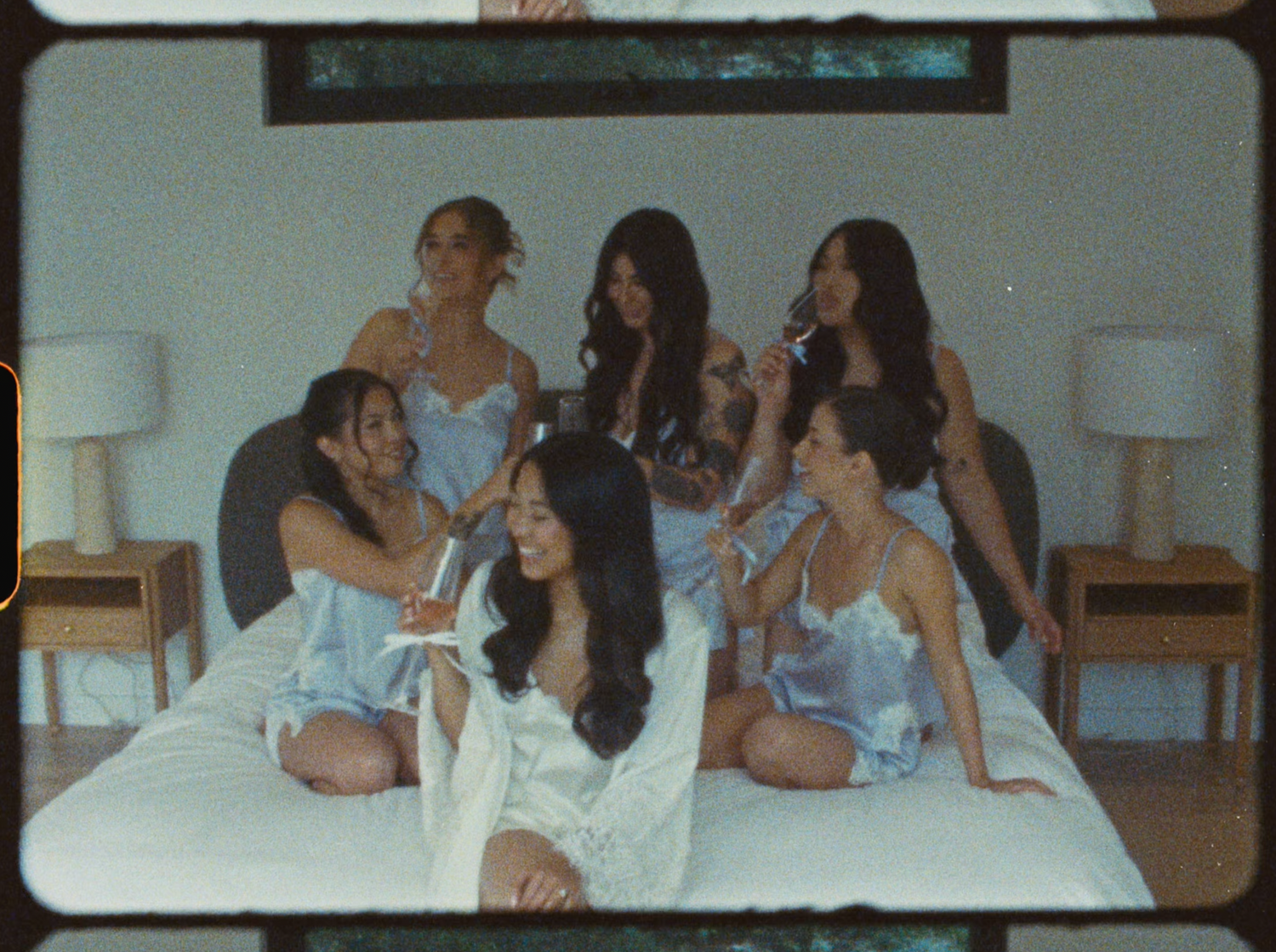 Six women in matching light blue satin camisoles gathered on a white bed celebrating together.