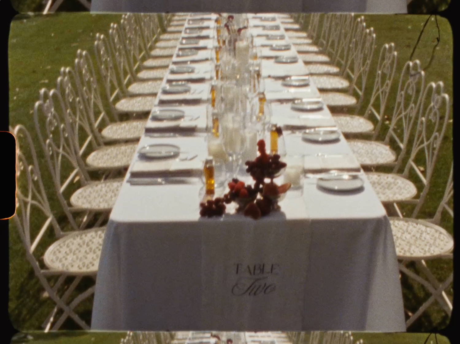 Long outdoor wedding banquet table with white linen labeled Table Three and burgundy floral centerpieces.