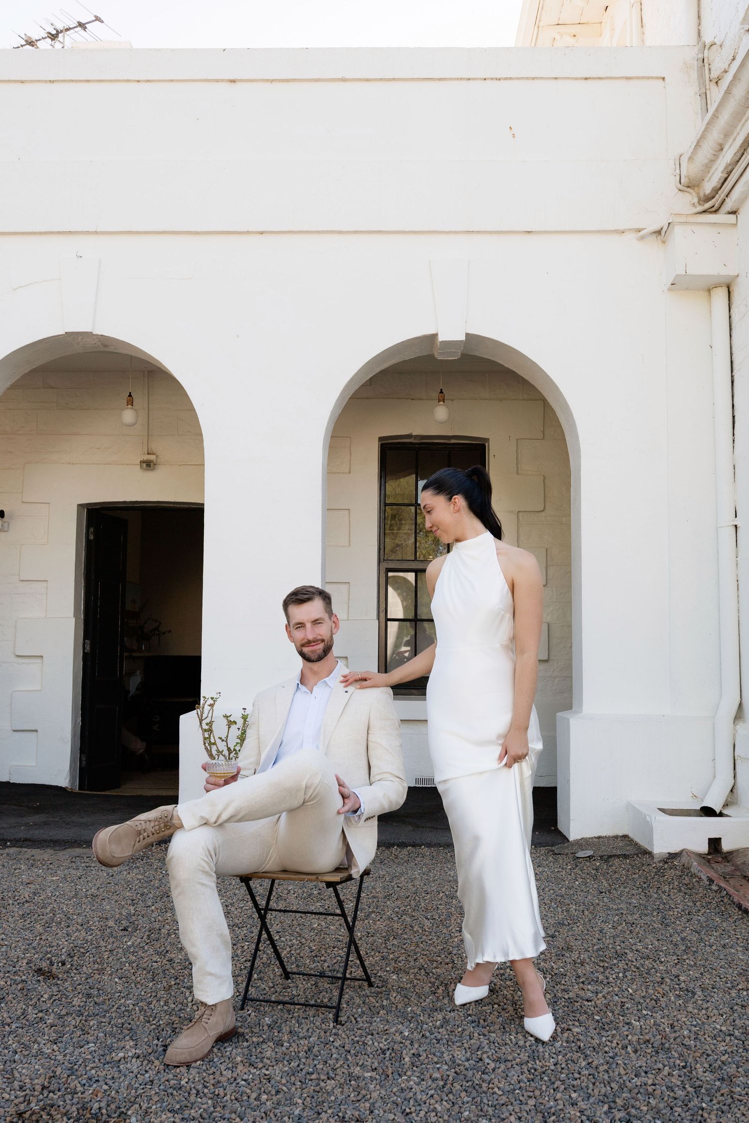 Groom seated outdoors smiles at bride in elegant white satin dress near white arches.