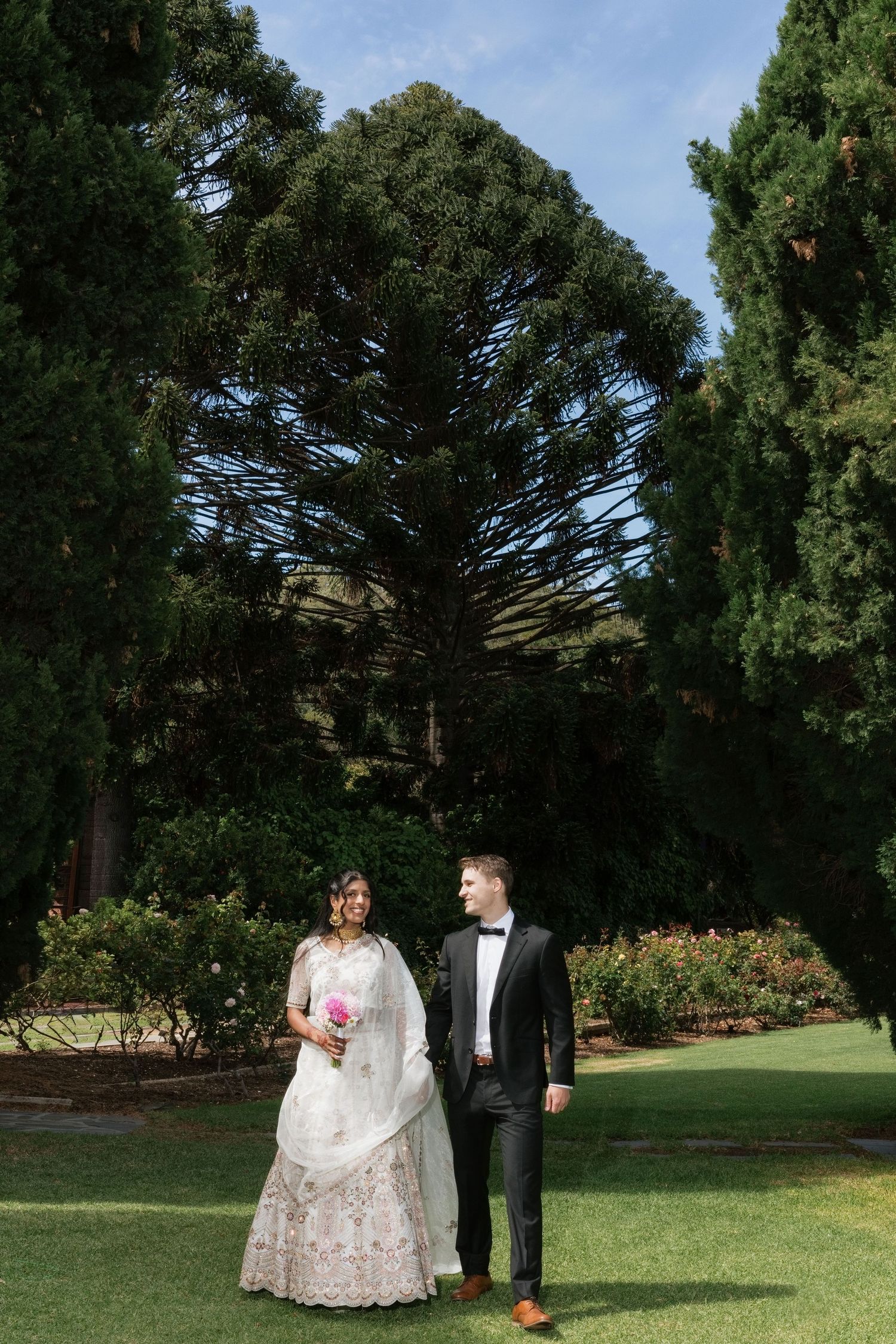 Bride and groom walking on green lawn, surrounded by tall trees, bride in white lehenga with bouquet.