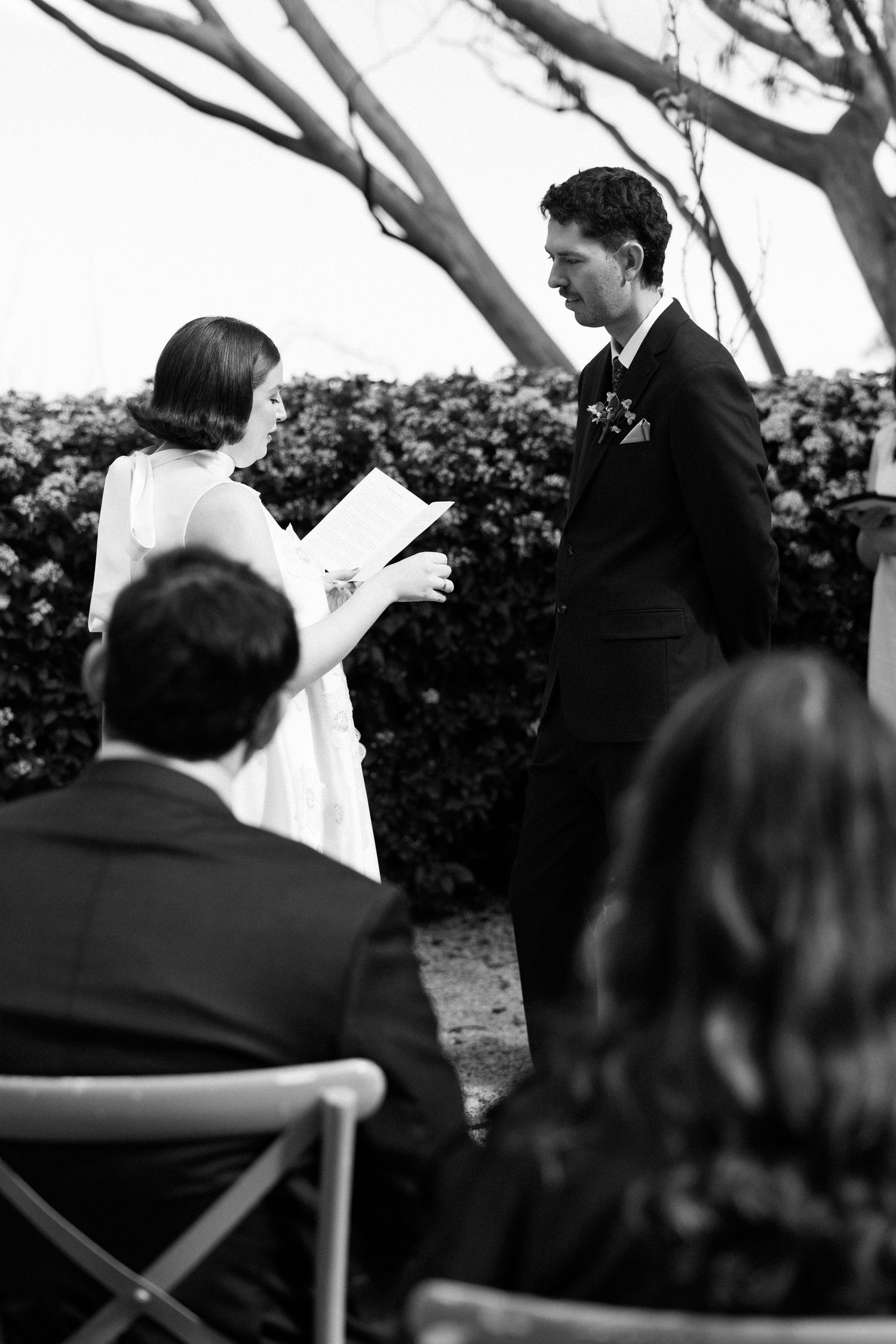 Black and white outdoor wedding ceremony with bride reading vows to groom, guests seated, floral hedge backdrop.