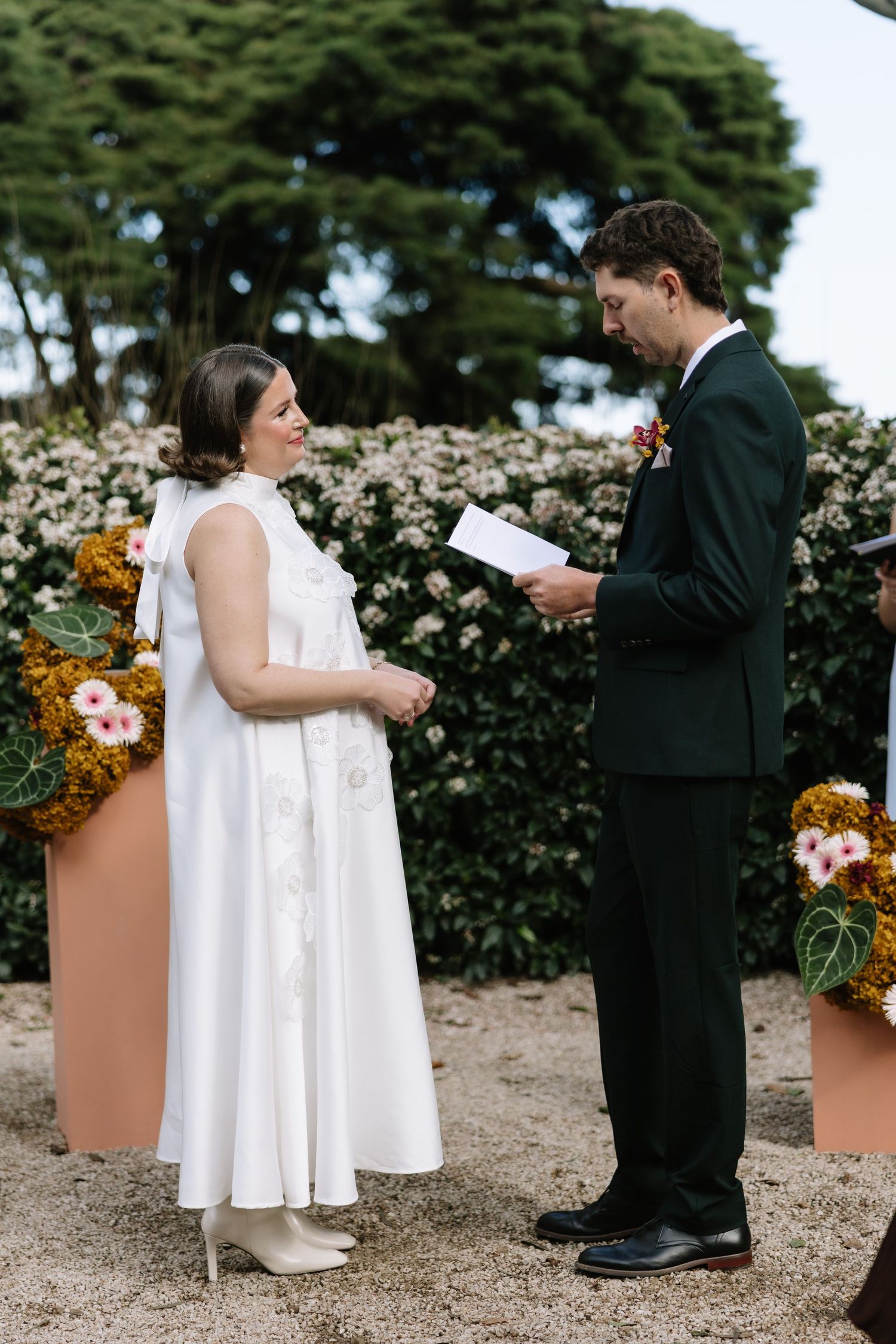 Bride and groom exchange vows outdoors, surrounded by lush greenery and floral arrangements at their wedding ceremony.