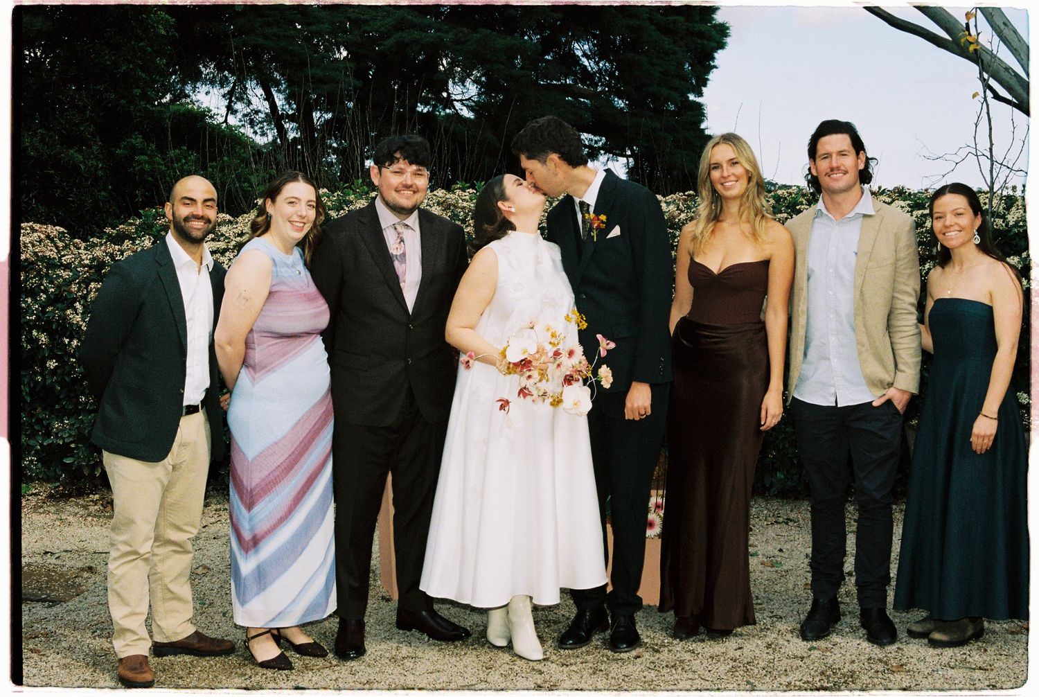 Wedding party group photo with bride and groom kissing, surrounded by six guests in formal attire outdoors.