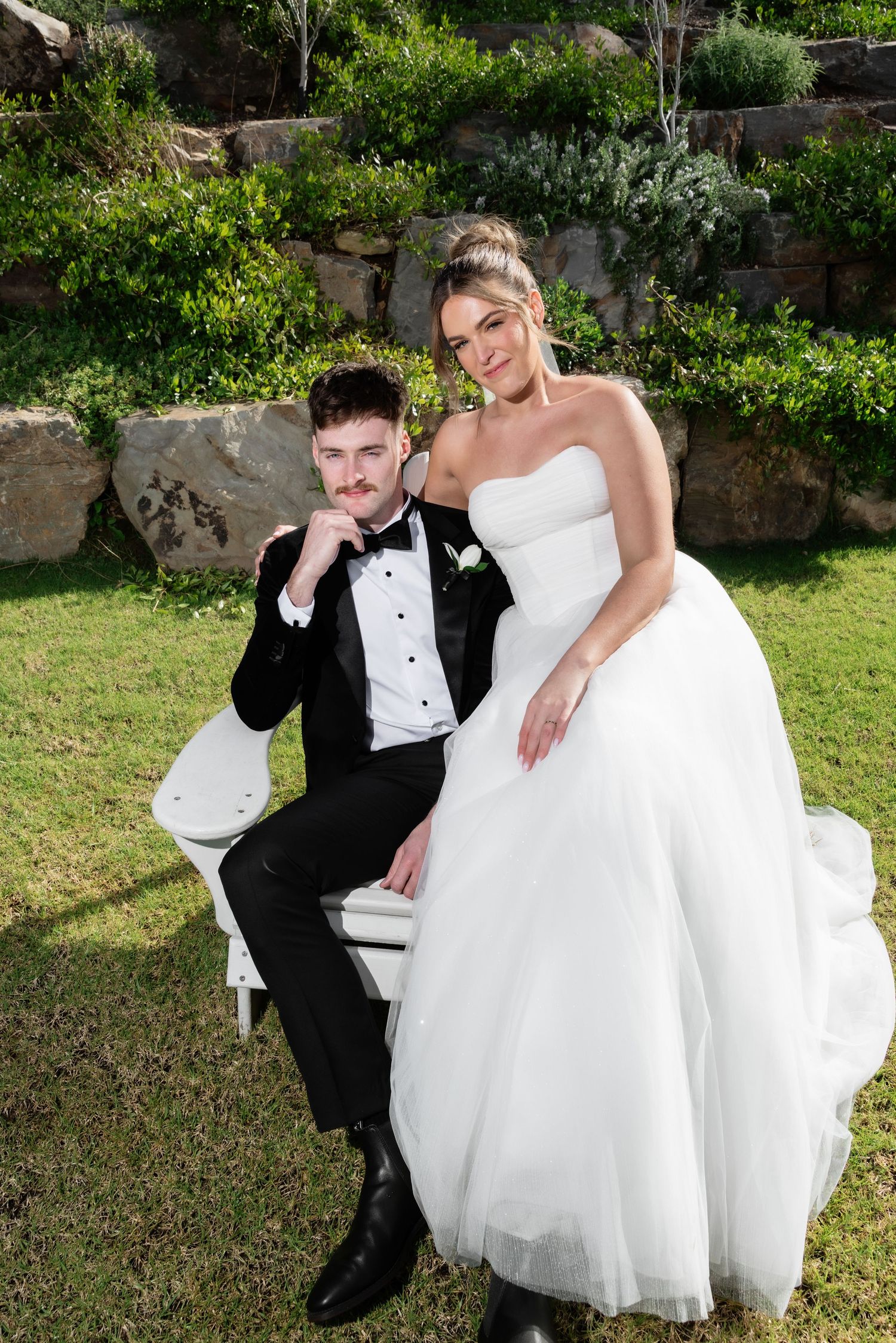 Bride in strapless white gown sits beside groom in black tuxedo on white bench in lush garden setting.