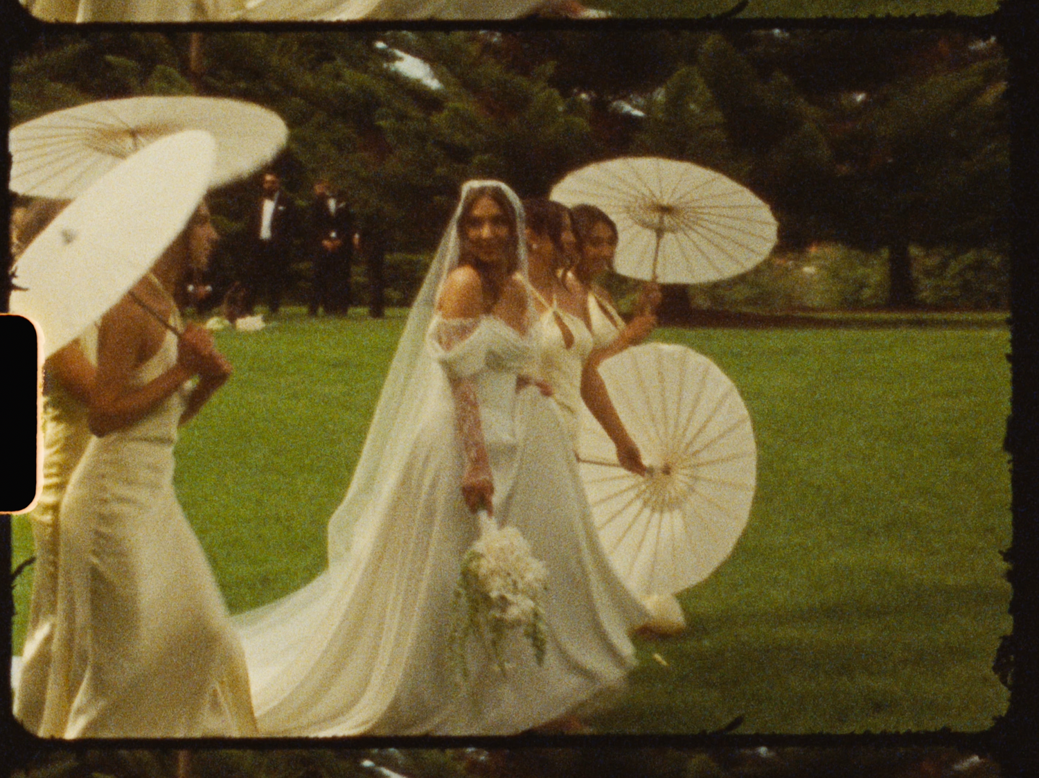 A bride in a white gown and veil walks outdoors with bridesmaids holding white parasols on a lush green lawn.