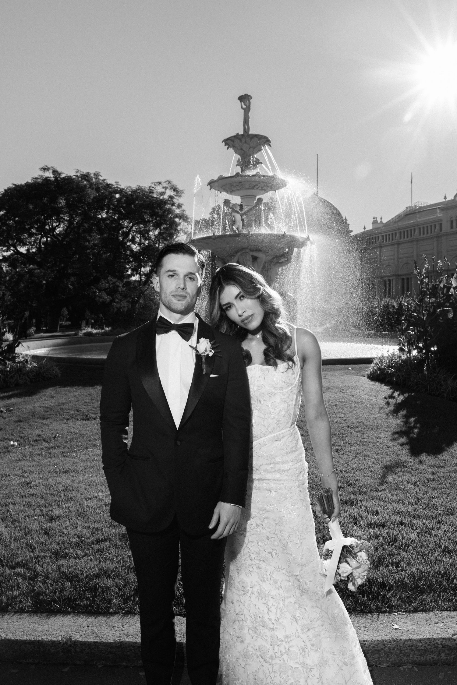 Black and white wedding photo of bride and groom posing before a sunlit fountain in a lush garden.