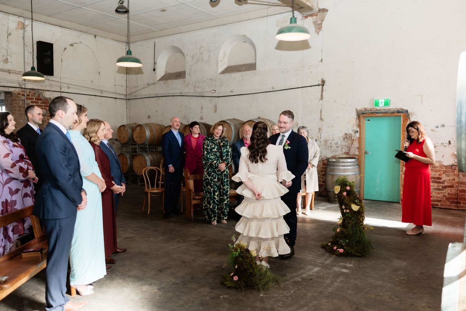 Wedding ceremony in a rustic barrel room with exposed brick, pendant lights, bride in ruffled gown and groom exchanging vows.