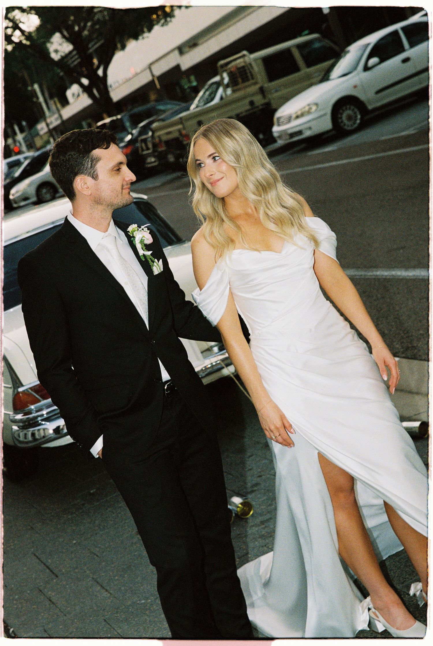 Stylish bride in off-shoulder white gown and groom in black suit posing beside vintage Mercedes.