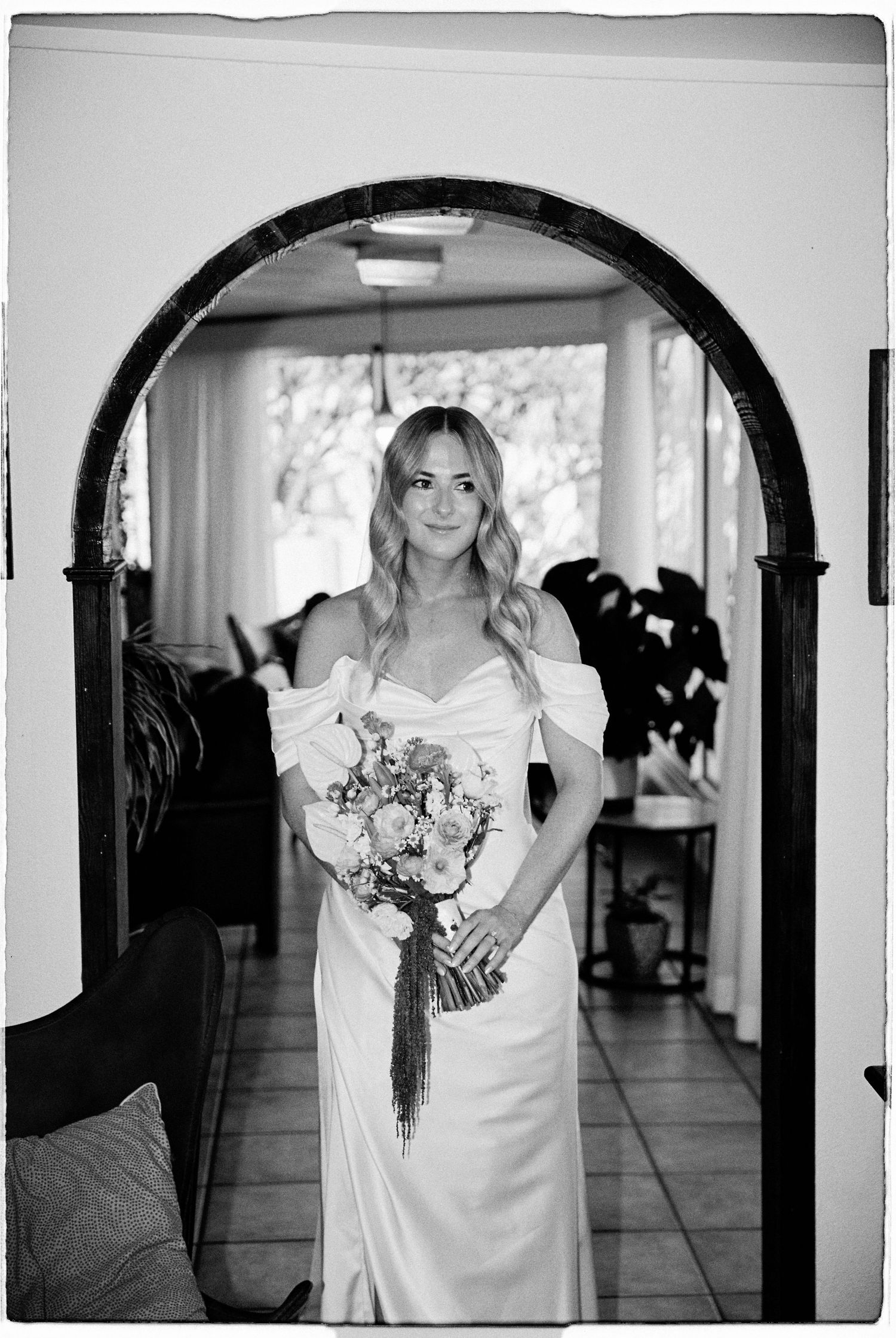 A bride in a white off-shoulder gown holding a bouquet under an arched doorway.
