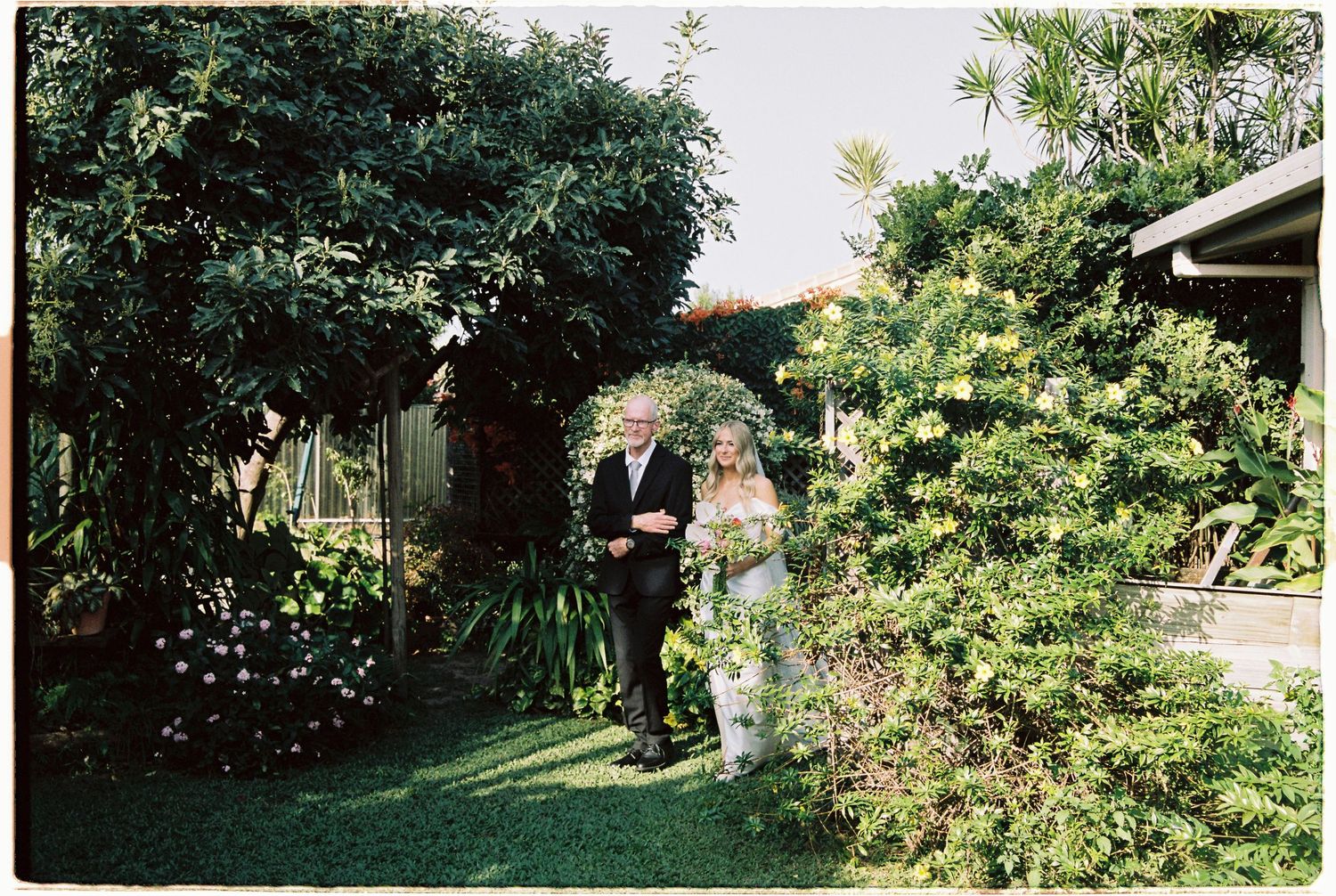 Bride walks through a beautiful garden pathway with escort during outdoor wedding ceremony.