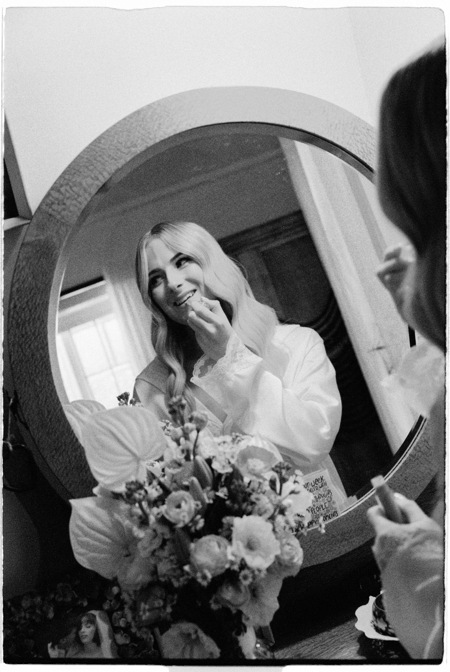 Black and white photo of a smiling blonde bride applying lipstick while looking into a round mirror with flowers.