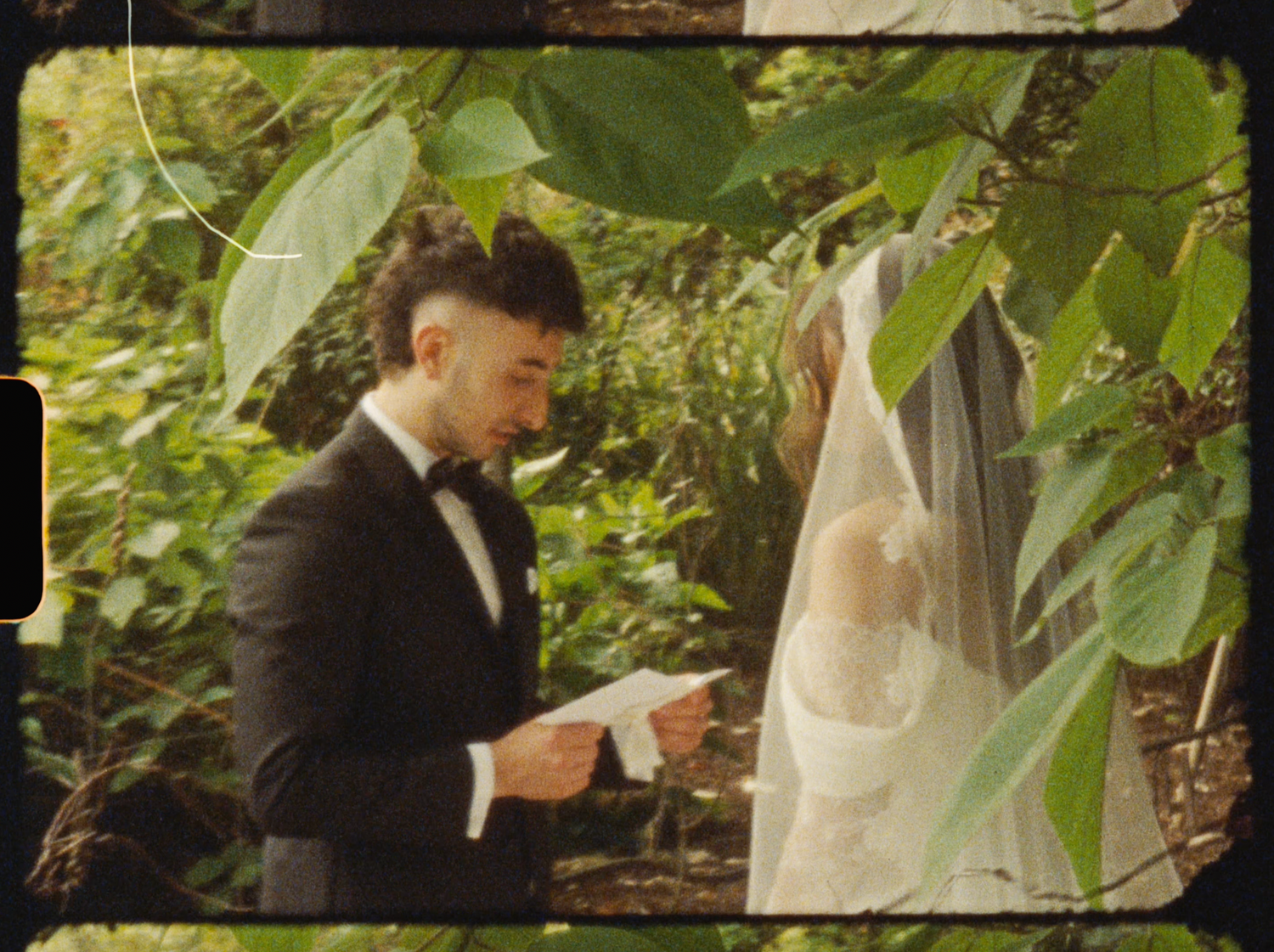 Groom in black tuxedo reads vows to bride in white gown and veil, surrounded by lush green garden foliage.