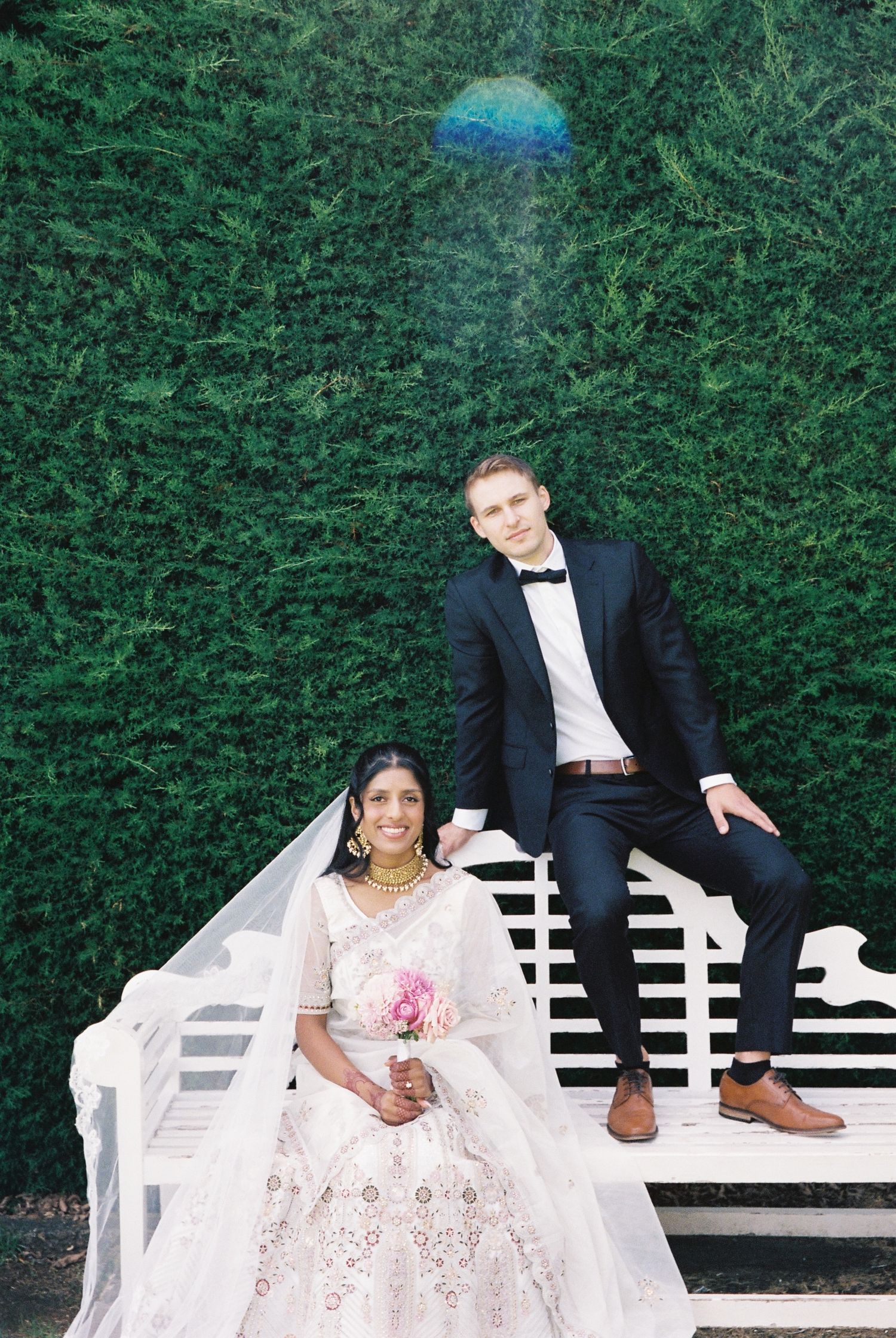 Bride in white lehenga and groom in black tuxedo posing on a white bench against a lush green hedge.