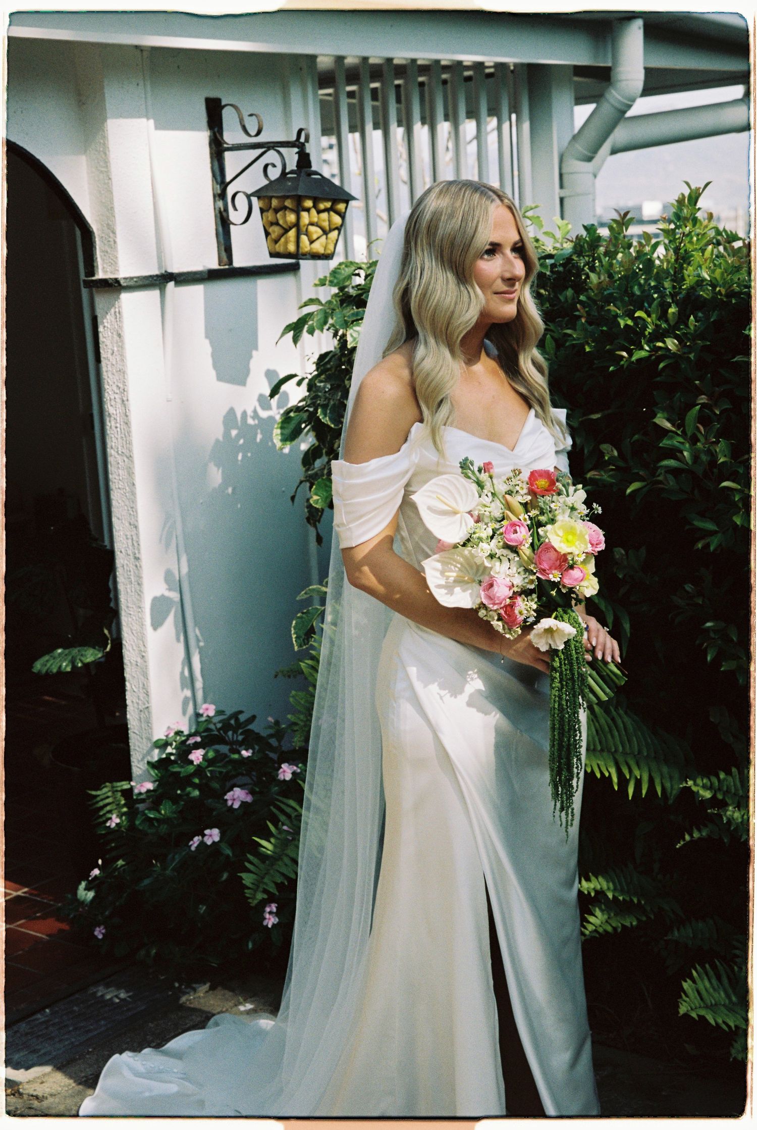 Bride in off-shoulder white gown with veil holds colorful bouquet outdoors near greenery and arched doorway.