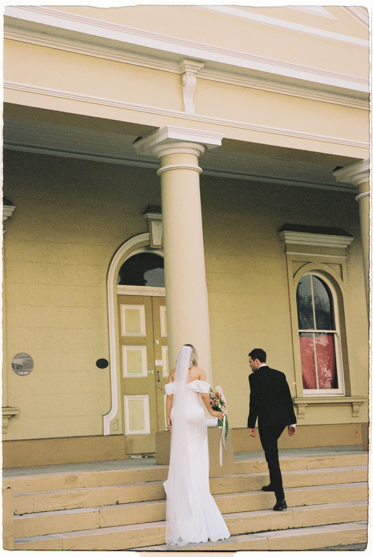 Bride in white off-shoulder gown and veil walks with groom in black tuxedo up steps of yellow historic building.