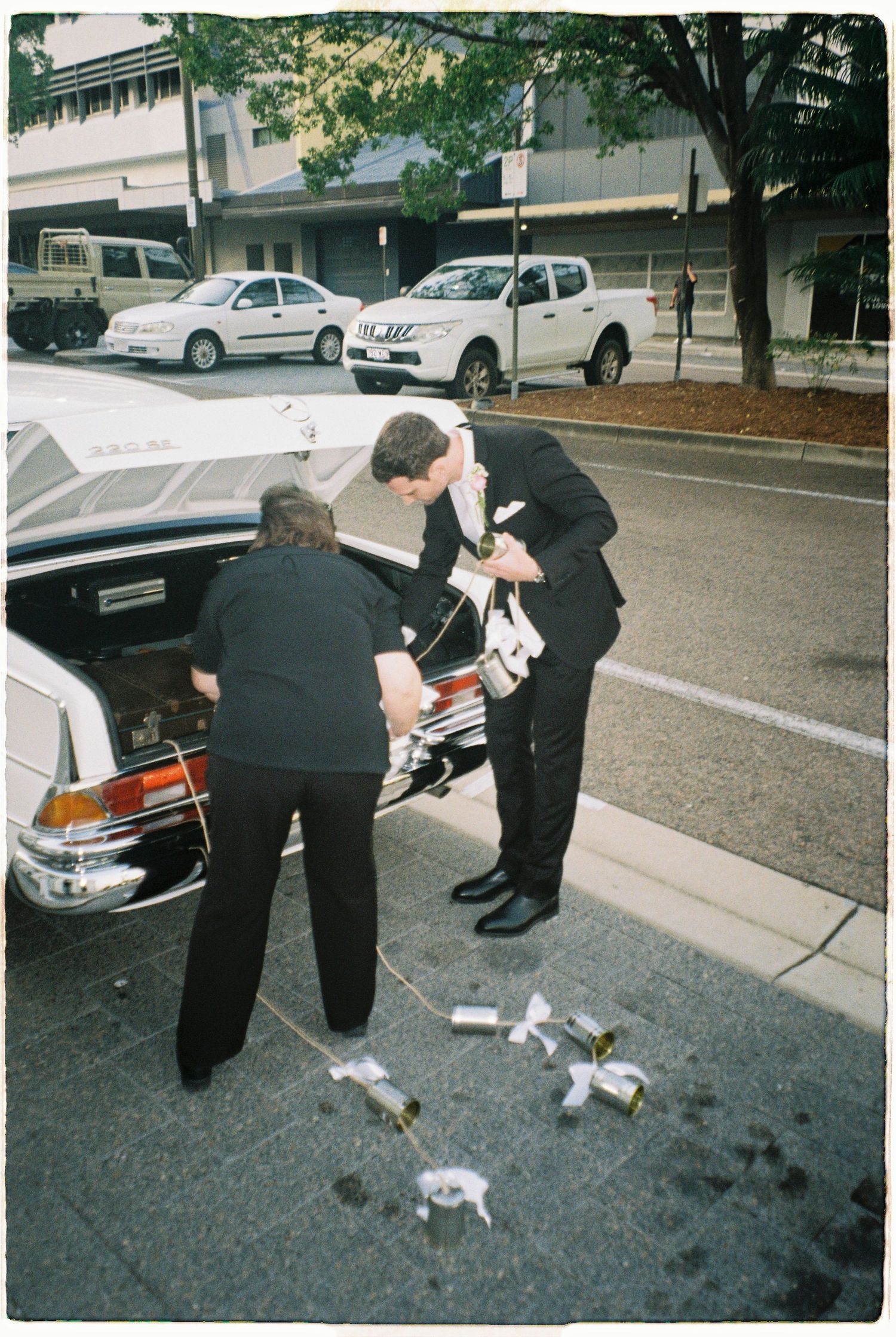 Groom in black suit retrieving items from the trunk of a vintage white wedding car on a city street.