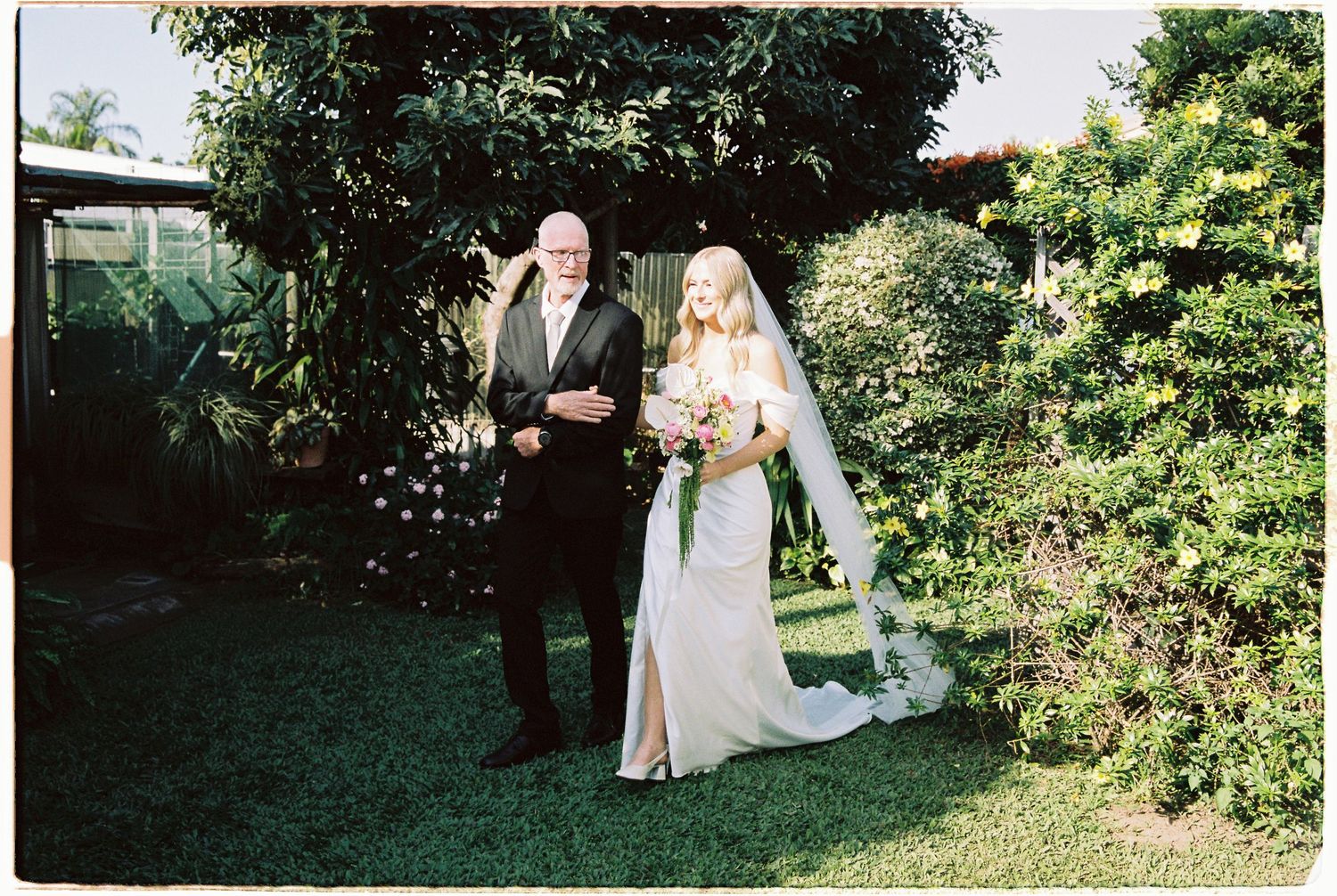 Bride in white gown and veil walks with older man in black suit through lush garden wedding venue.