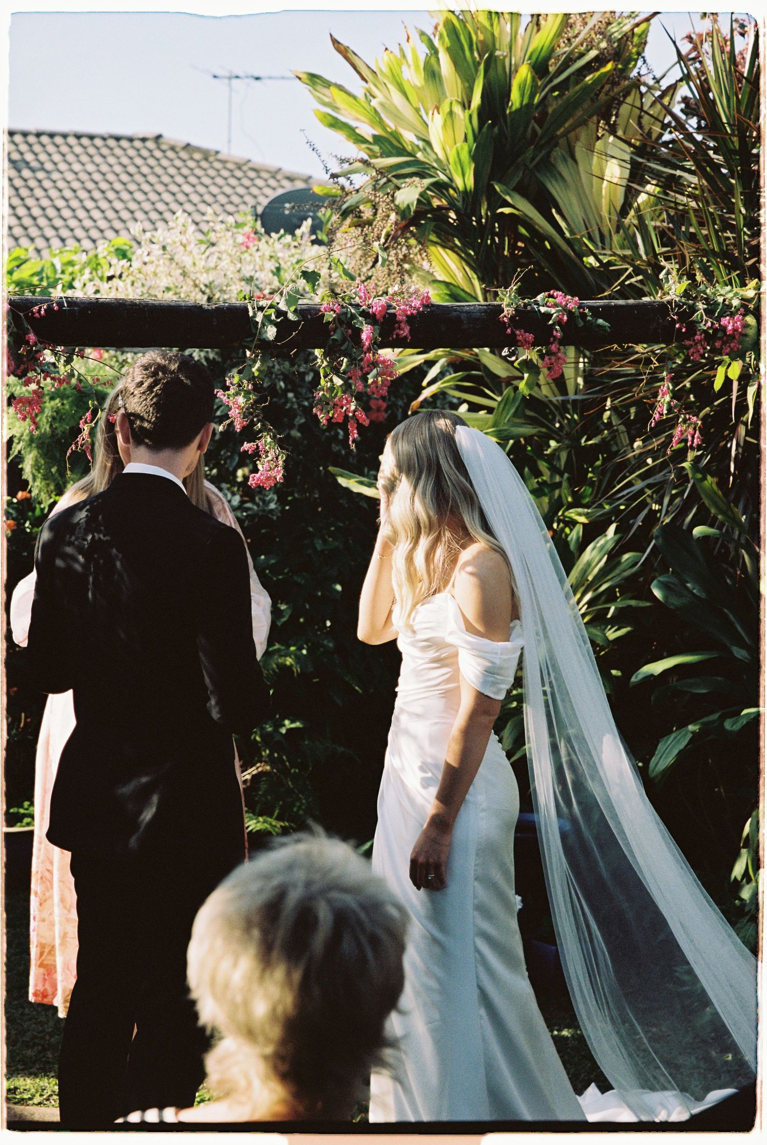 Bride in white gown and veil stands with groom under floral garden arch.