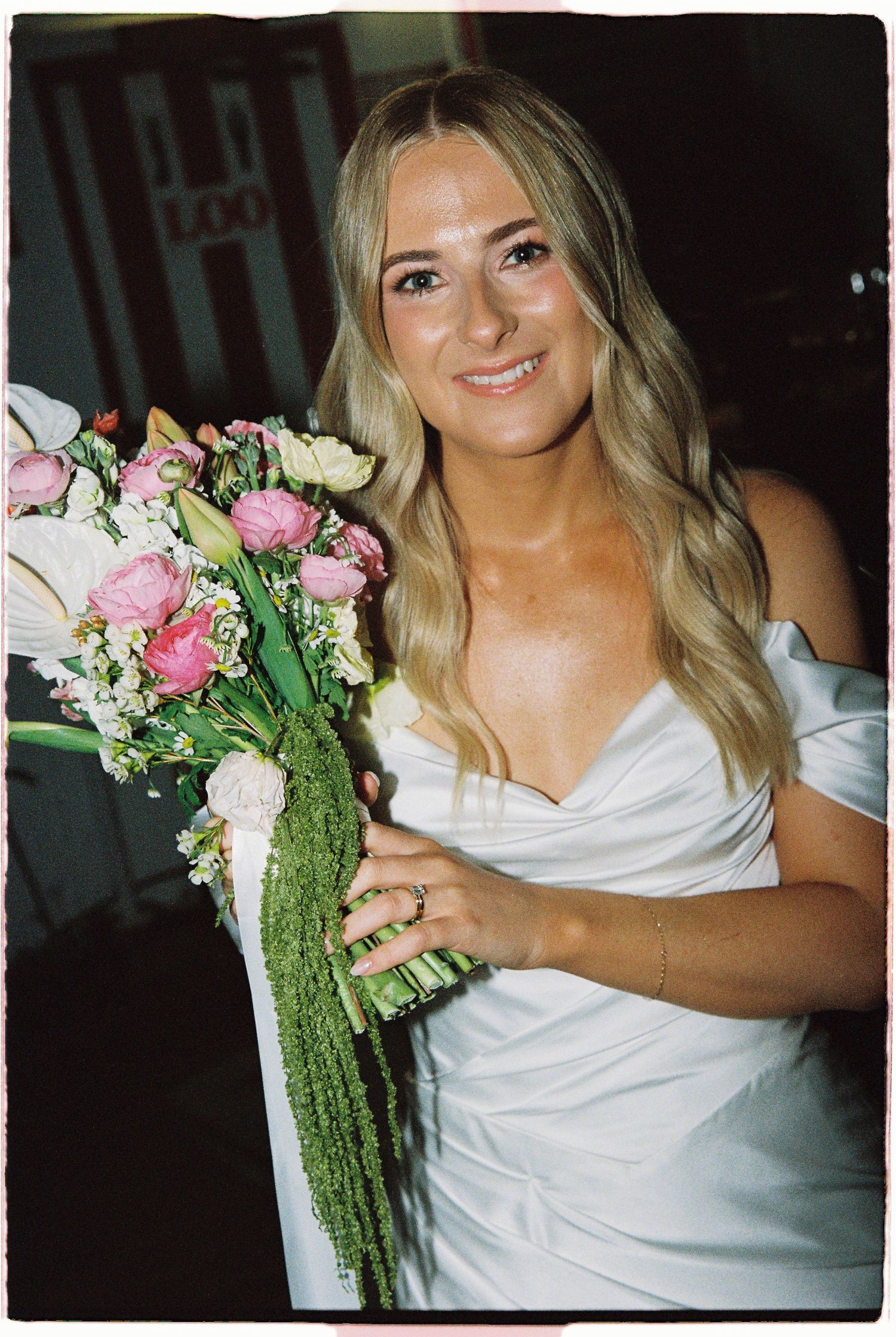 Smiling bride in white off-shoulder dress holding a cascading pink and white floral bouquet.
