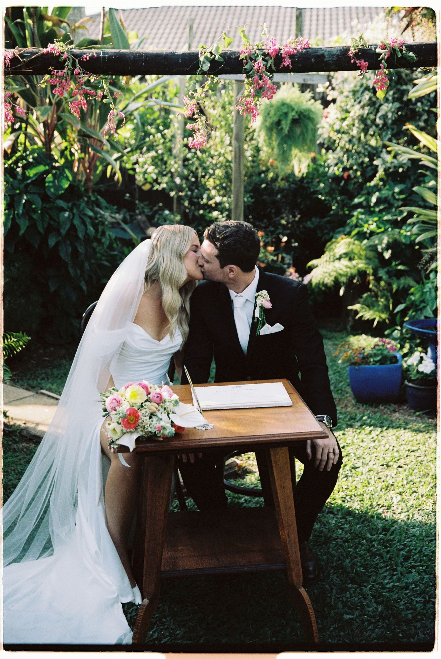 Bride and groom kiss at wooden signing table beneath hanging floral arch in sunlit garden ceremony.