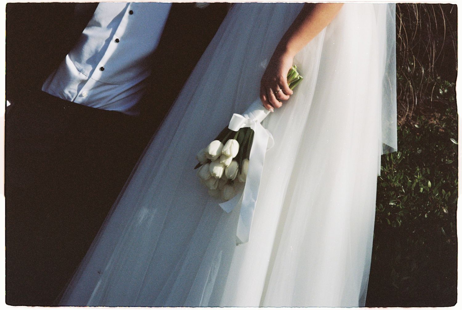 Bride in white wedding gown holding a small white tulip bouquet beside groom outdoors.