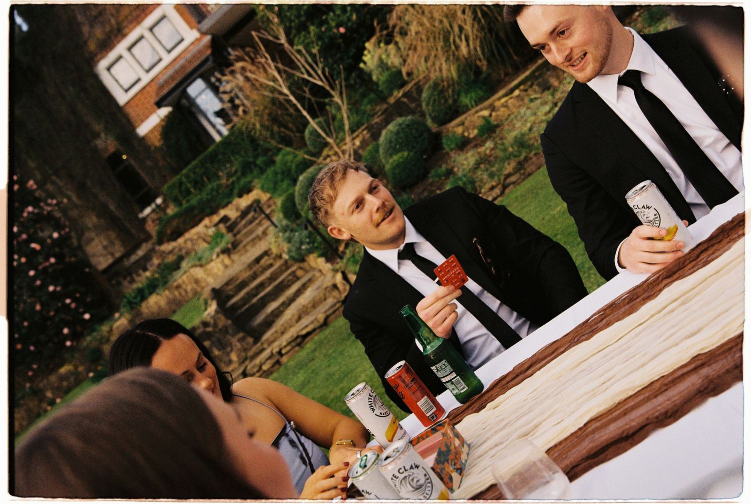 Wedding guests in formal attire socializing at an outdoor reception table with drinks and bottles.