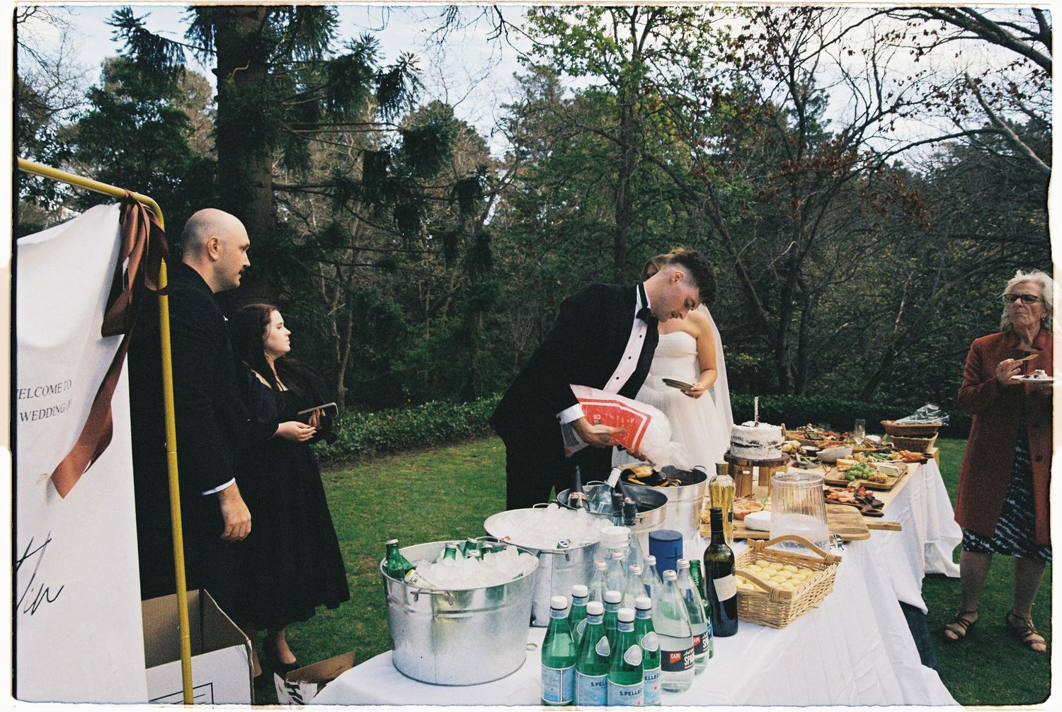 Outdoor wedding reception buffet table with guests mingling in a lush garden setting.