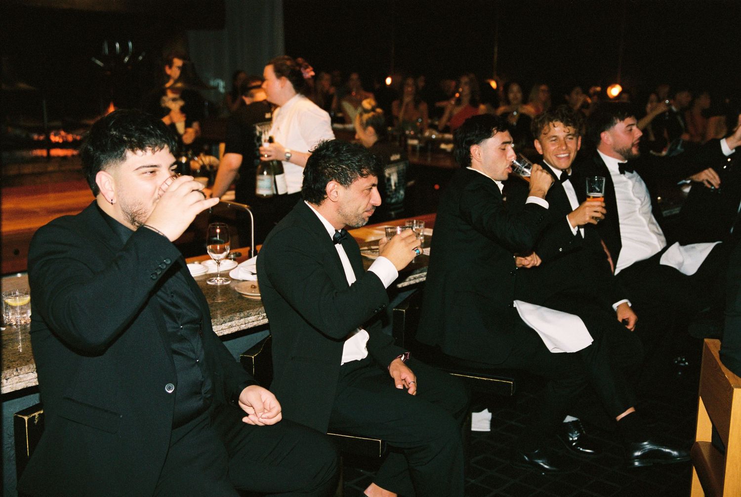 A group of men in black tuxedos laugh and drink together while seated at a wedding reception.